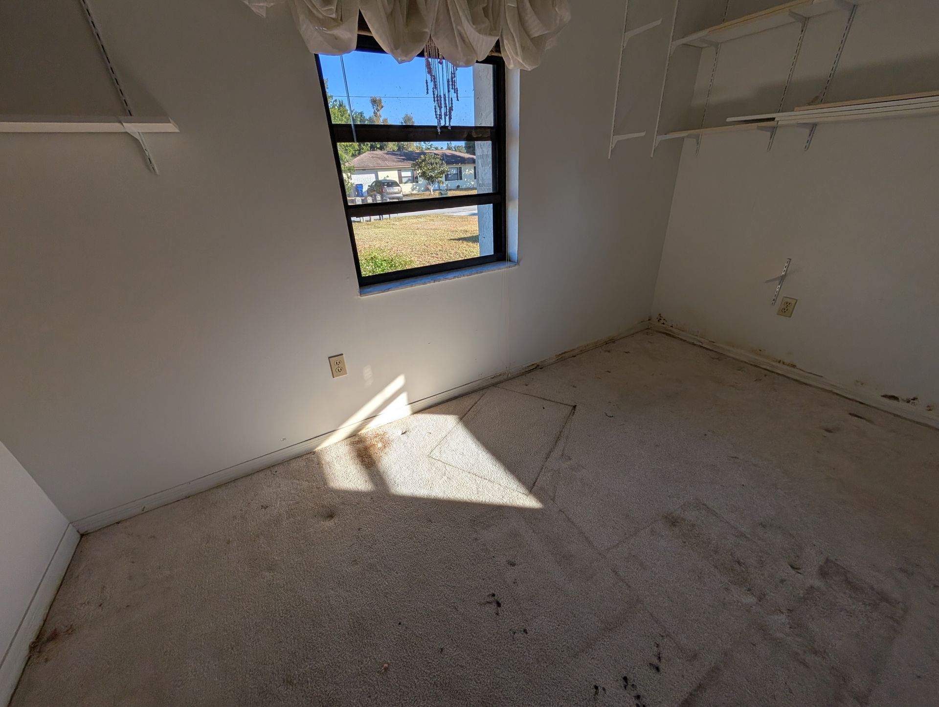 Small, empty room with stained carpet, window, and shelves. Sunlight shines in.