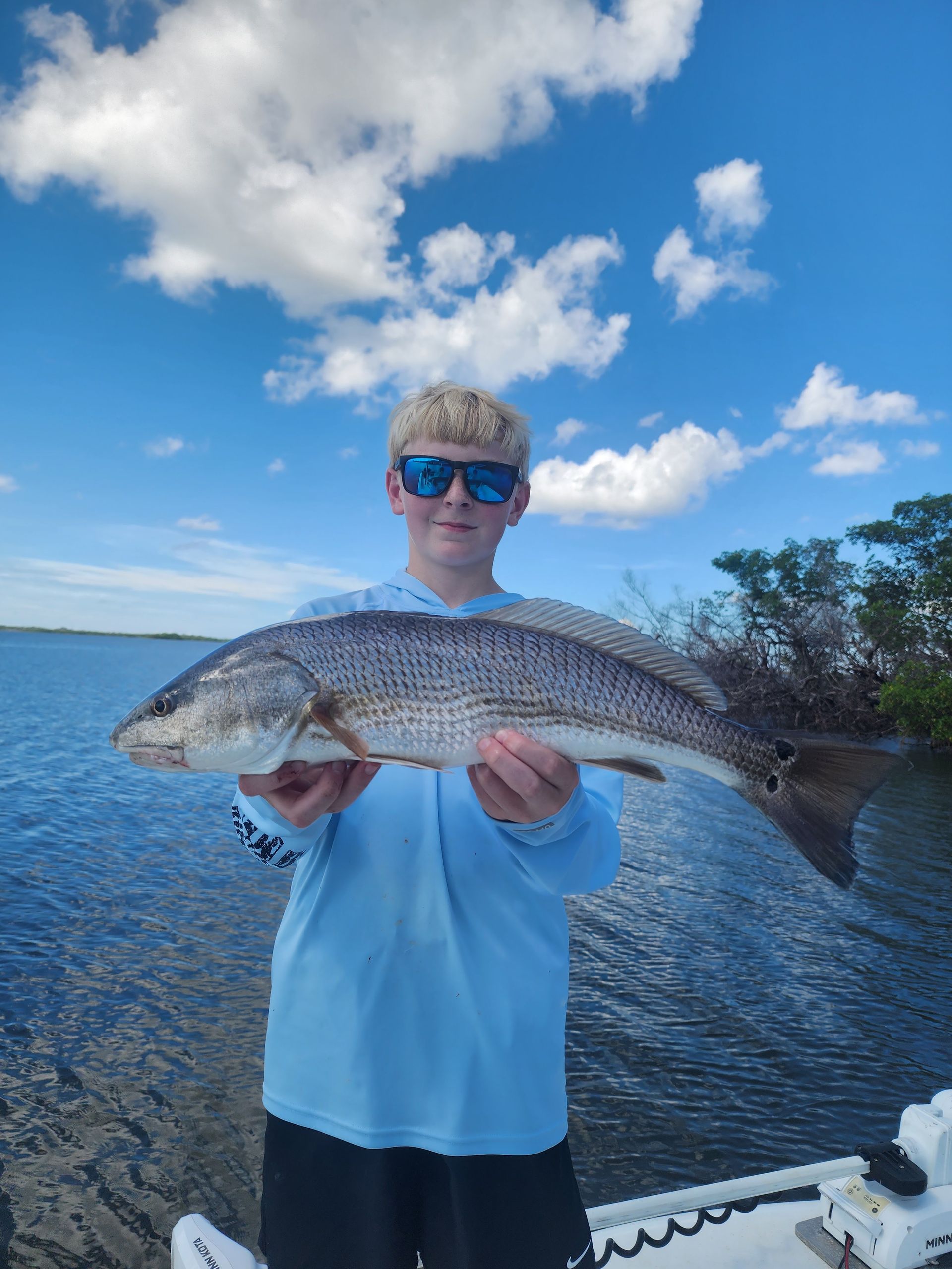 A young man is holding a large fish in his hands in front of a body of water.