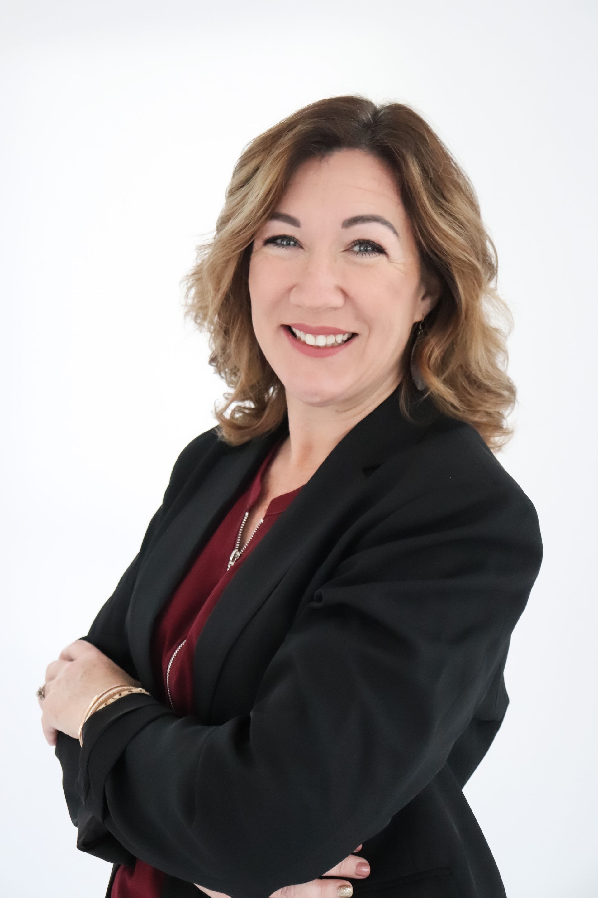 A woman in a black jacket and red shirt is smiling with her arms crossed.