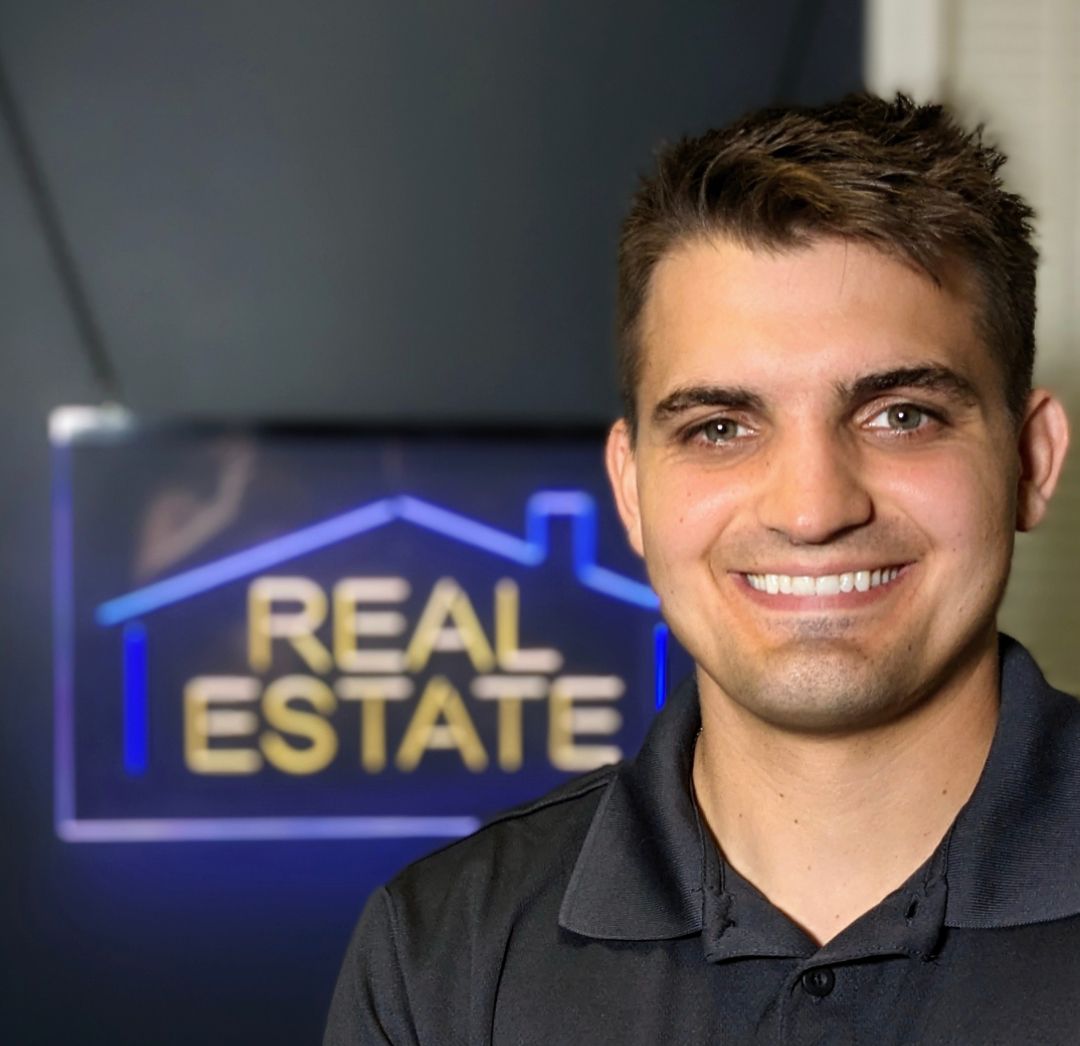A man is smiling in front of a real estate sign