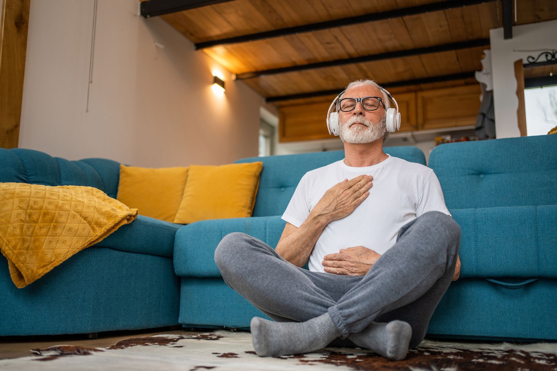 An elderly man is sitting on the floor in front of a couch wearing headphones.