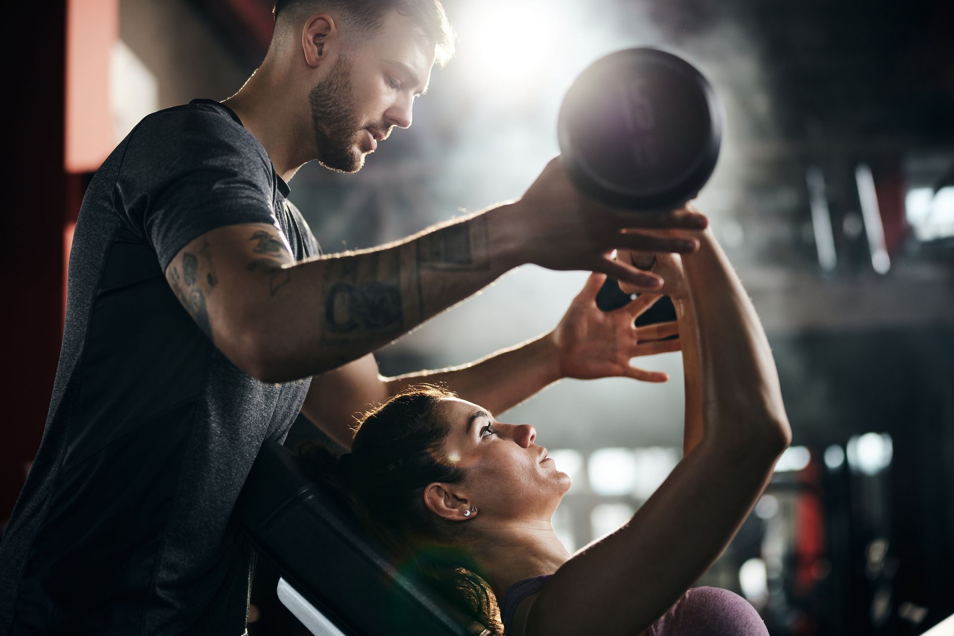 A man is helping a woman lift a medicine ball in a gym.