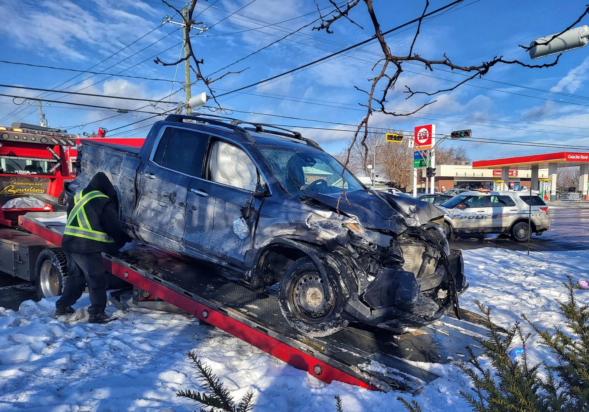 Une camionnette sombre, lourdement endommagée, est remorquée après une collision sur une route enneigée. Un dépanneur se tient à proximité, avec une voiture de police en arrière-plan.