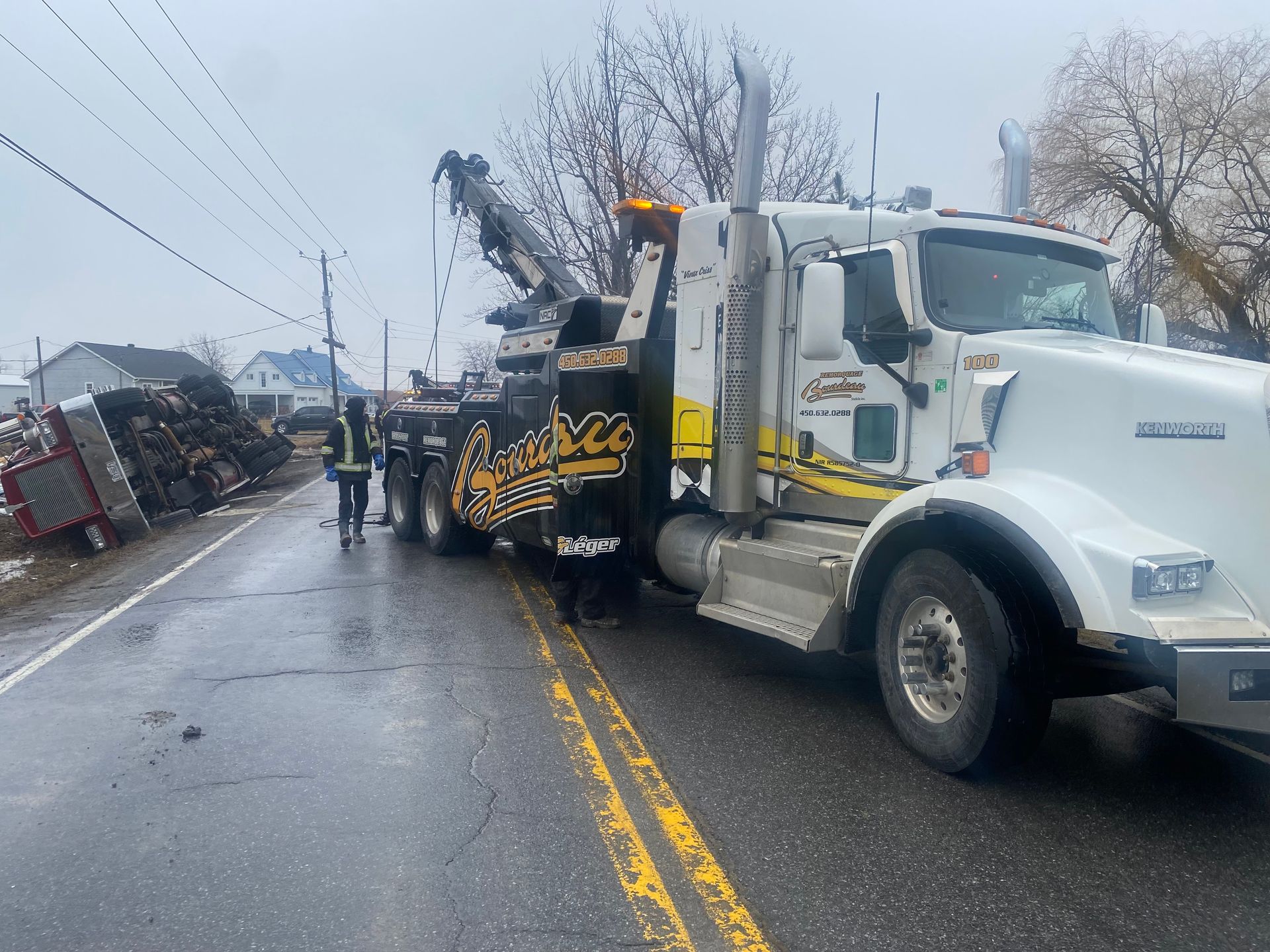 Une dépanneuse sur les lieux d'un accident impliquant une remorque à plateau renversée et chargée de métal. Deux personnes portant des gilets pare-balles se tiennent près du lieu de l'accident, sur une route mouillée.