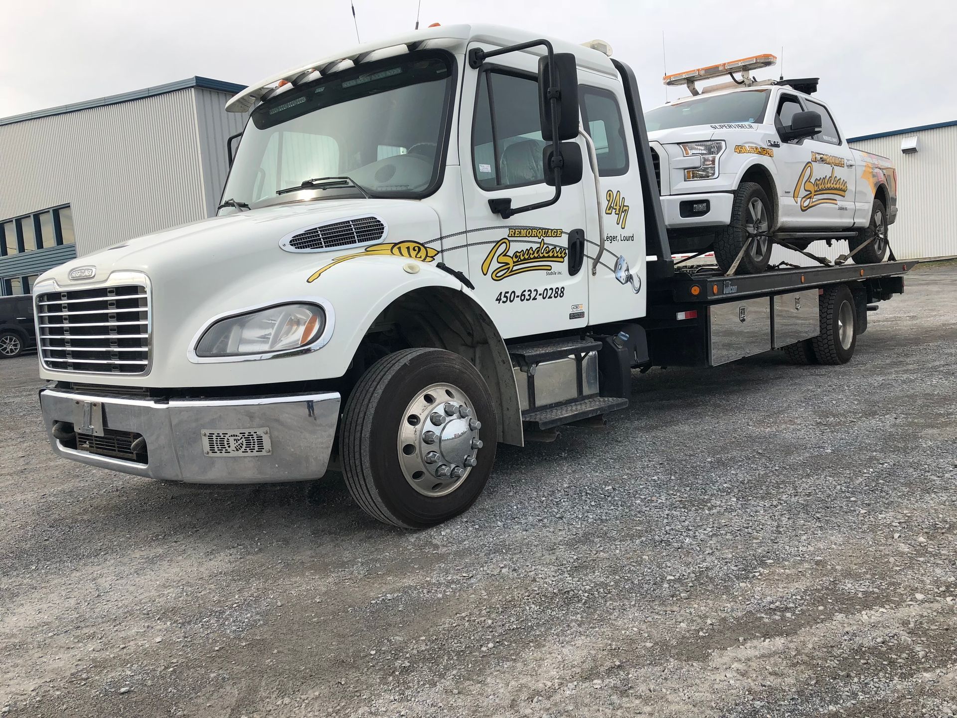 Une dépanneuse blanche avec une voiture chargée sur son plateau, garée dehors. Les deux véhicules portent le logo de l'entreprise.