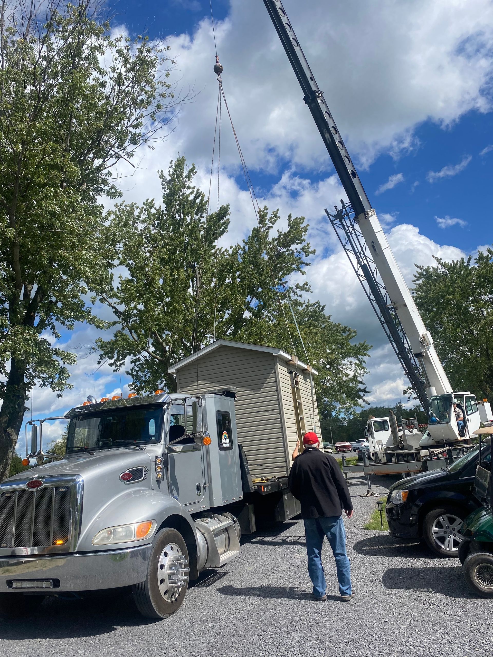 Un hangar est soulevé d'un camion par une grue. Une personne portant une casquette rouge observe. Le ciel bleu et les arbres sont en arrière-plan.