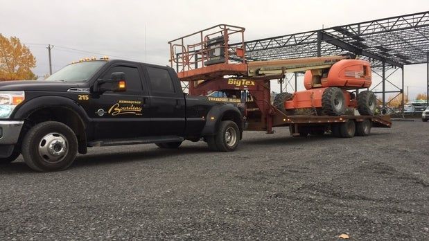 Camionnette noire tractant une nacelle élévatrice orange sur une remorque dans un terrain en gravier sous un ciel couvert.