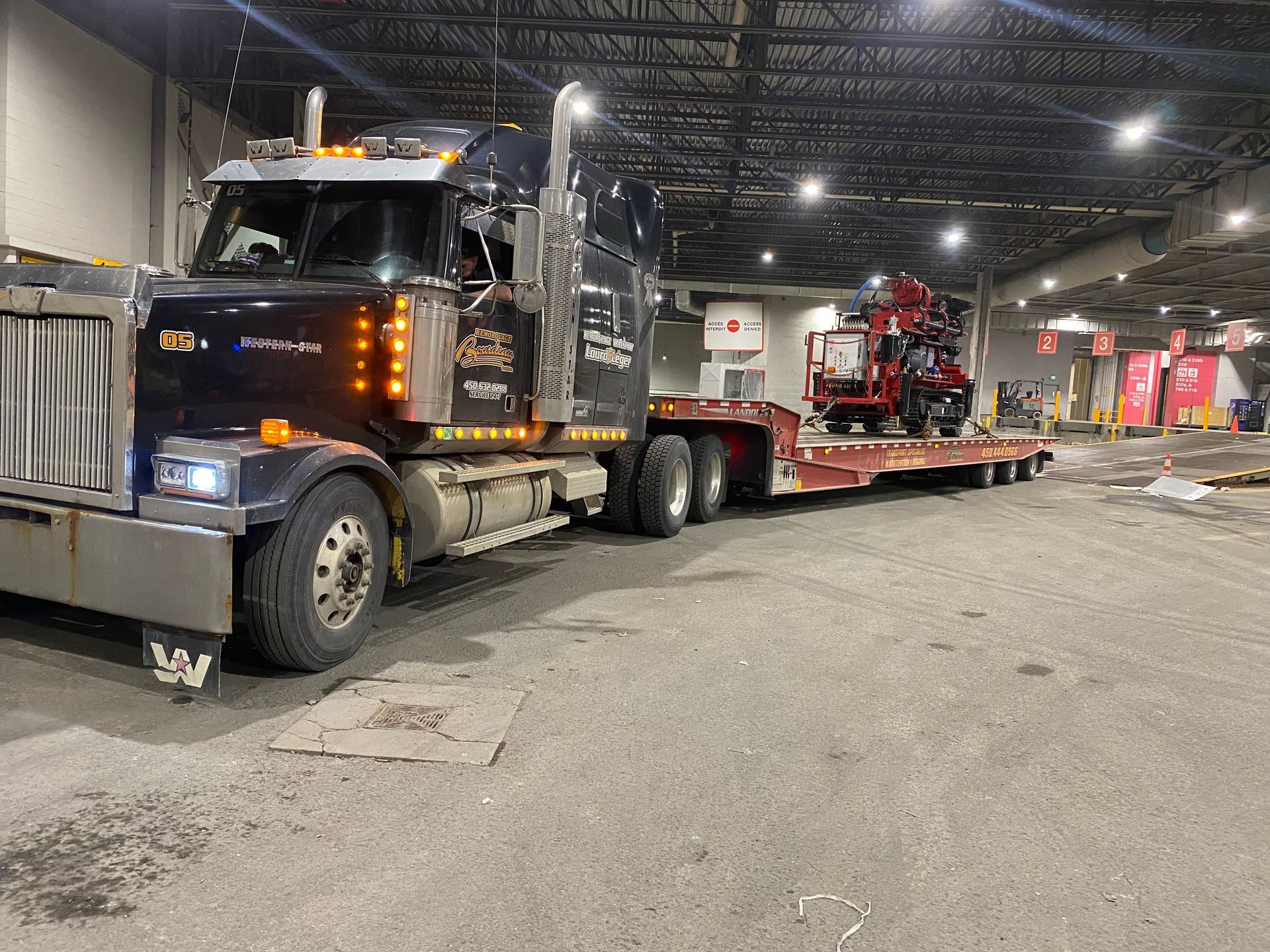 Semi-remorque noir avec une remorque surbaissée transportant une machine agricole rouge, garée à l'intérieur d'un entrepôt.
