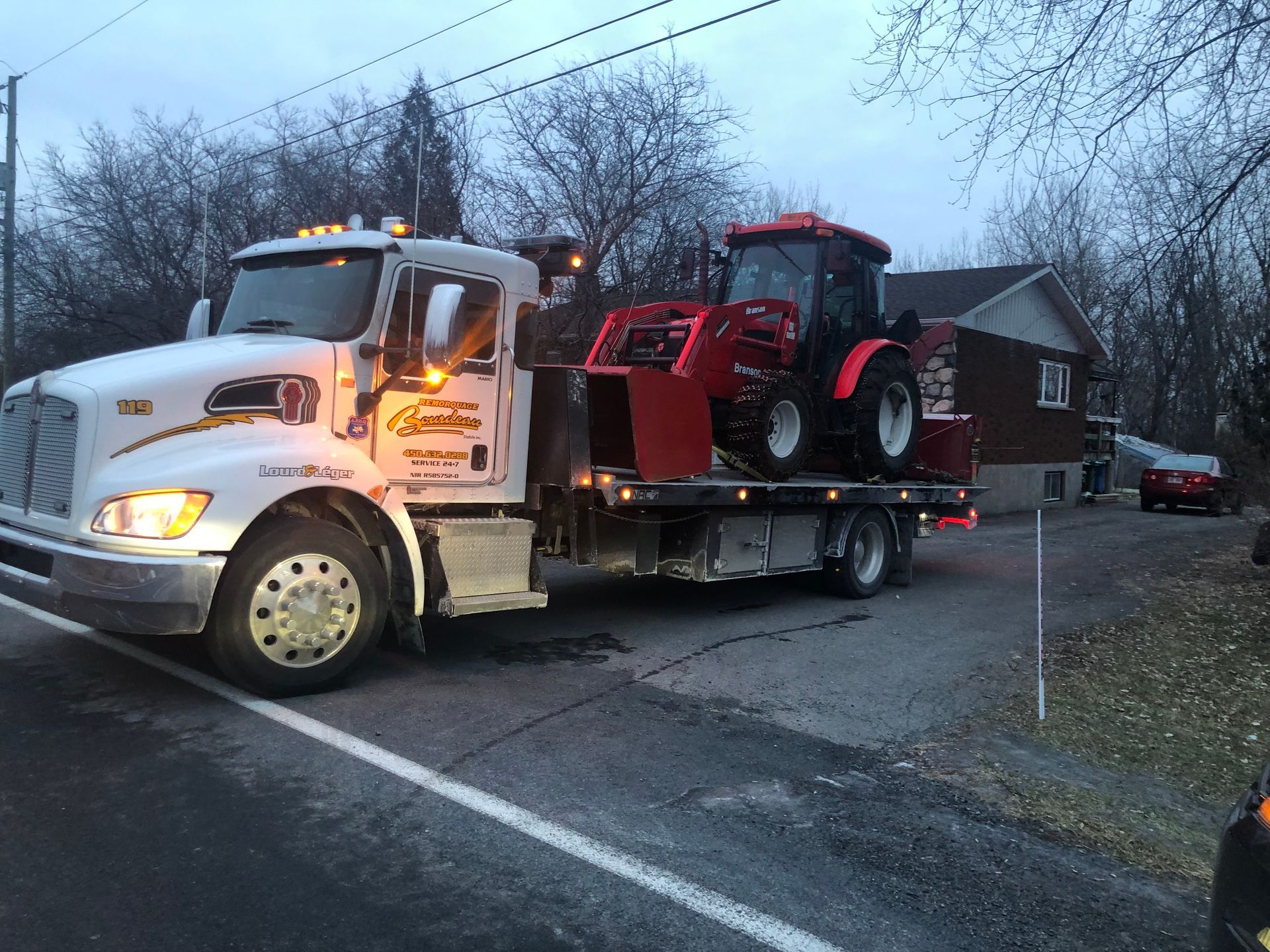 Une dépanneuse à plateau transportant un tracteur rouge est garée sur une route près d'une maison. Le ciel est couvert.