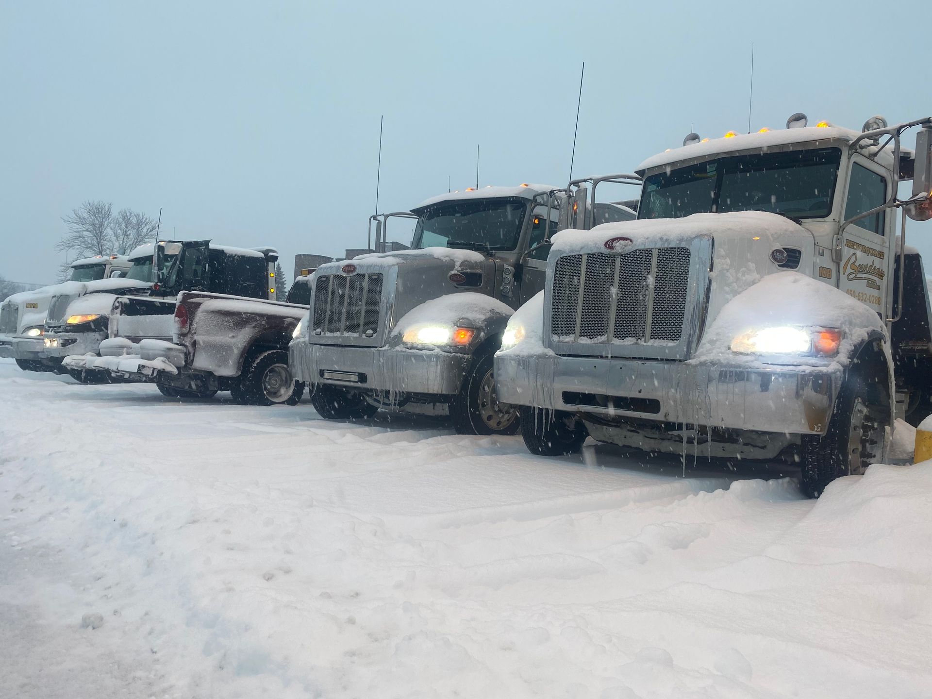 Des camions enneigés sont garés sur un parking enneigé. Des véhicules gris aux phares brillants font face au spectateur, la neige tombant dessus.