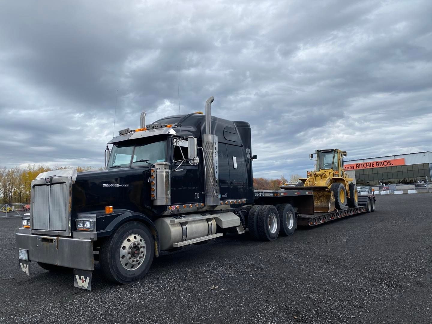 Semi-camion noir transportant une chargeuse de chantier jaune sur une remorque à plateau dans un parking sous un ciel nuageux.