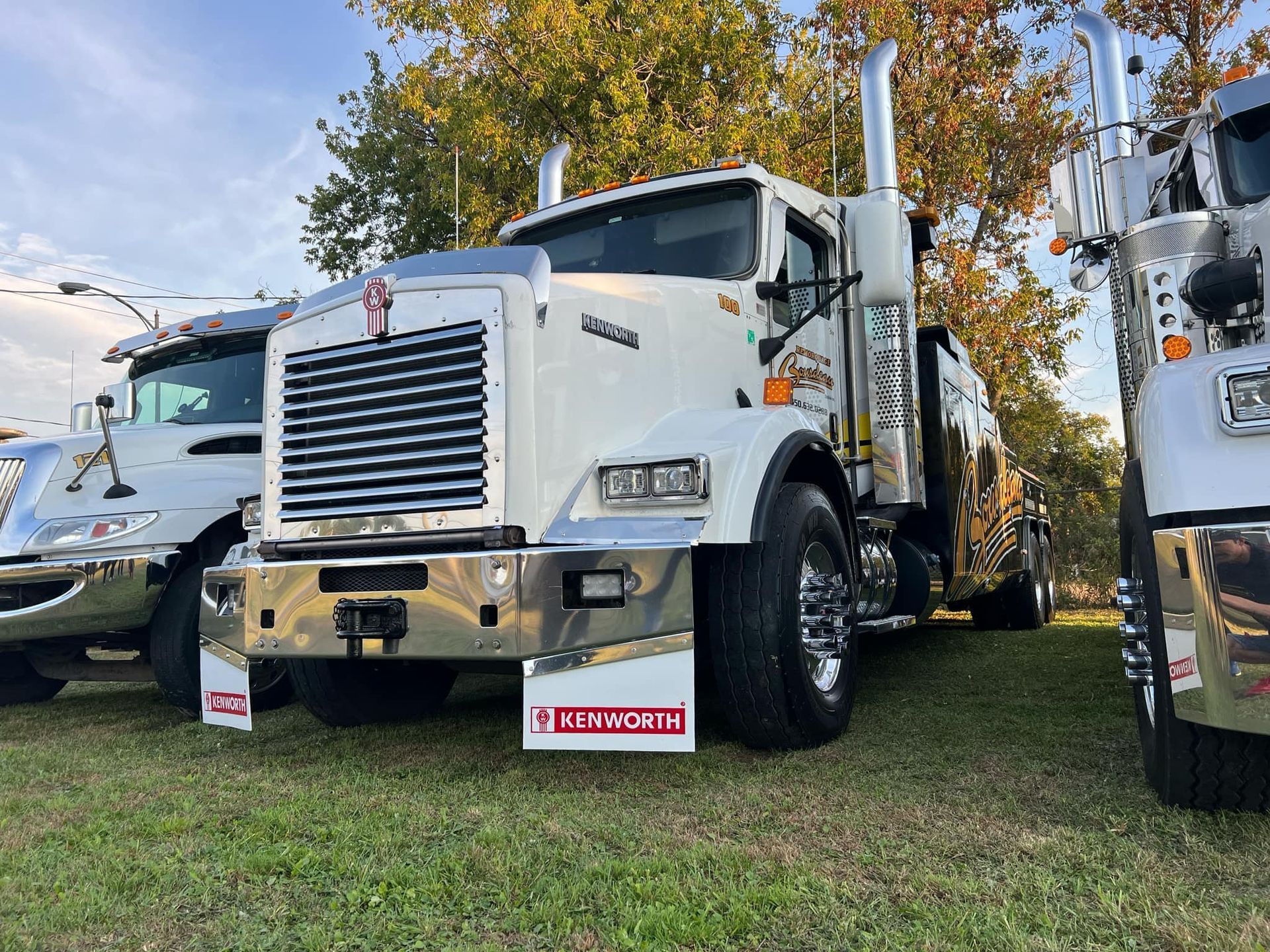 Dépanneuse Kenworth blanche garée sur l'herbe, vue de face. Journée ensoleillée. D'autres camions sont visibles à proximité.