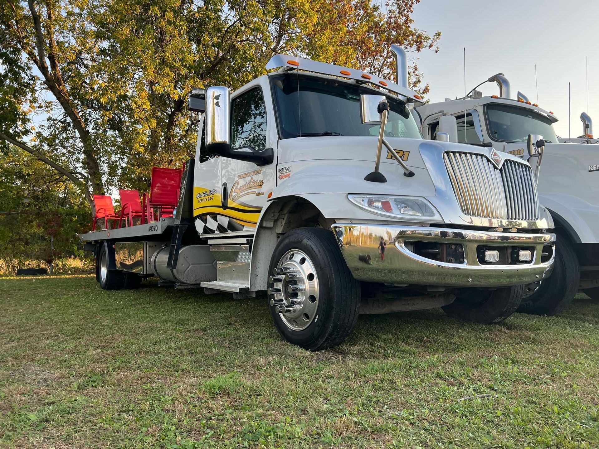 Dépanneuse blanche avec plateau garée sur l'herbe, avec une vue partielle d'un autre camion en arrière-plan.