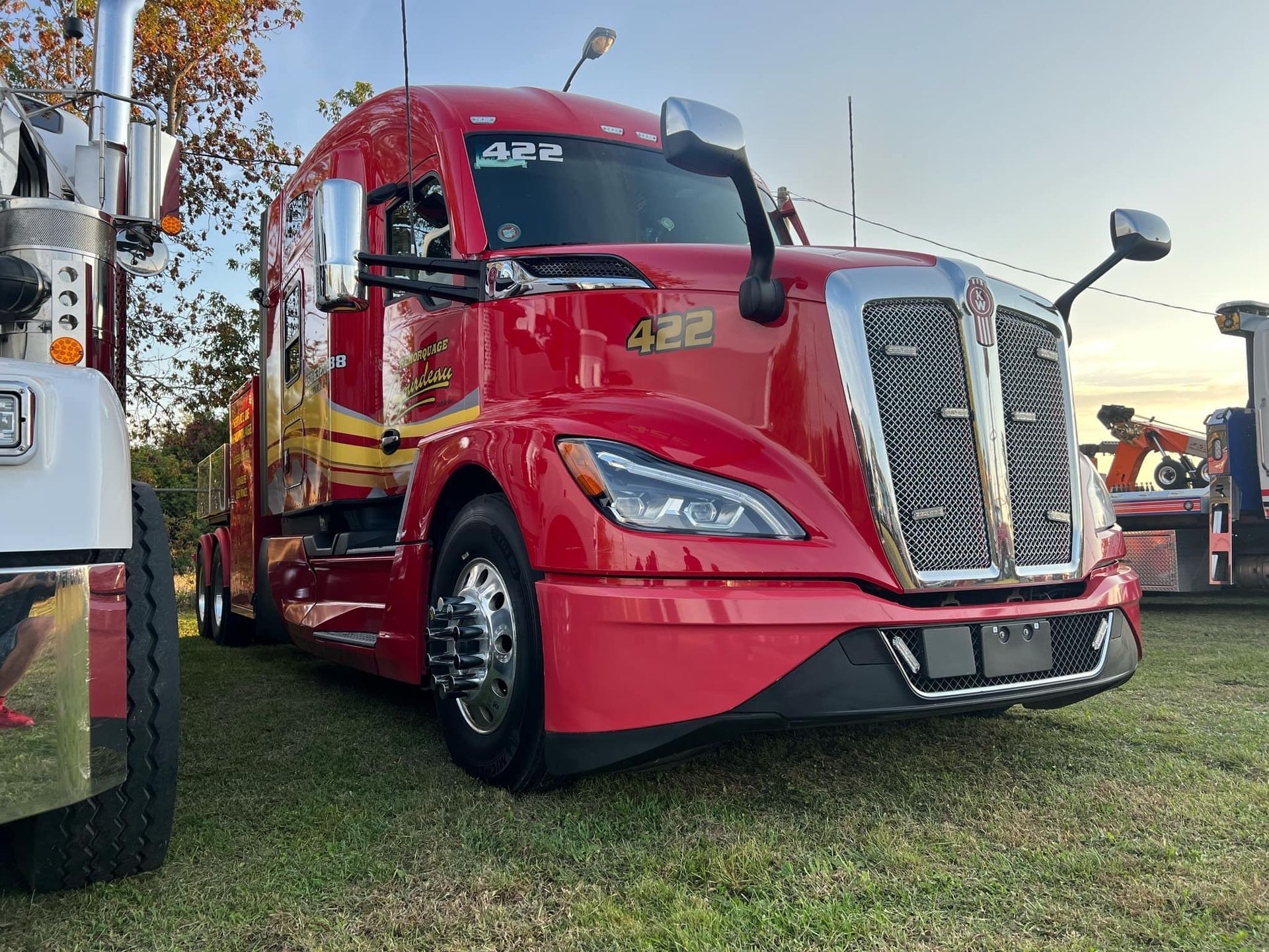 Semi-remorque Kenworth rouge garée sur l'herbe, mettant en valeur sa calandre et ses rétroviseurs. Le décor semble être en extérieur.