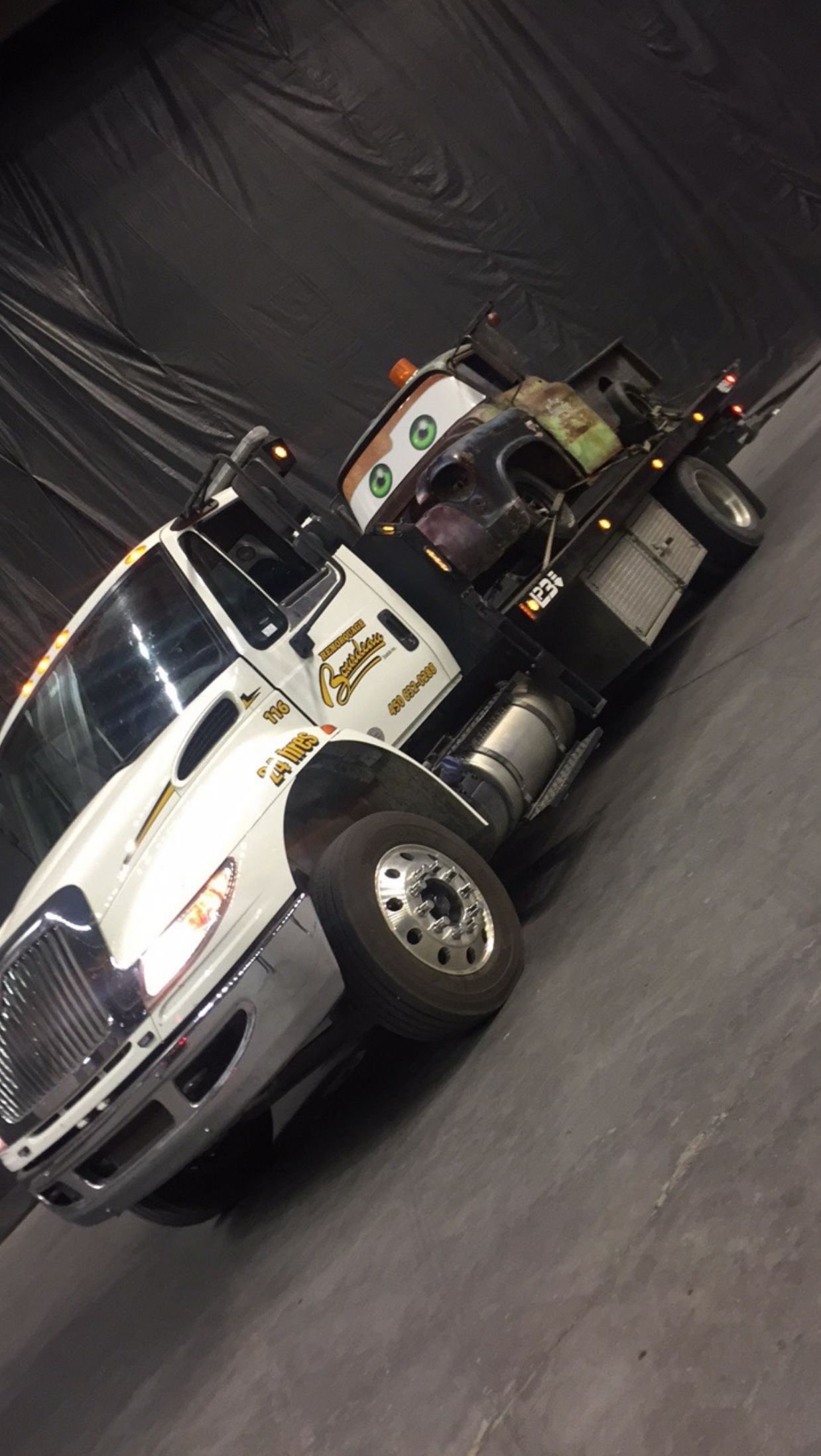 Dépanneuse blanche sur sol gris, vue en angle. Son bras est relevé, prêt à remorquer.