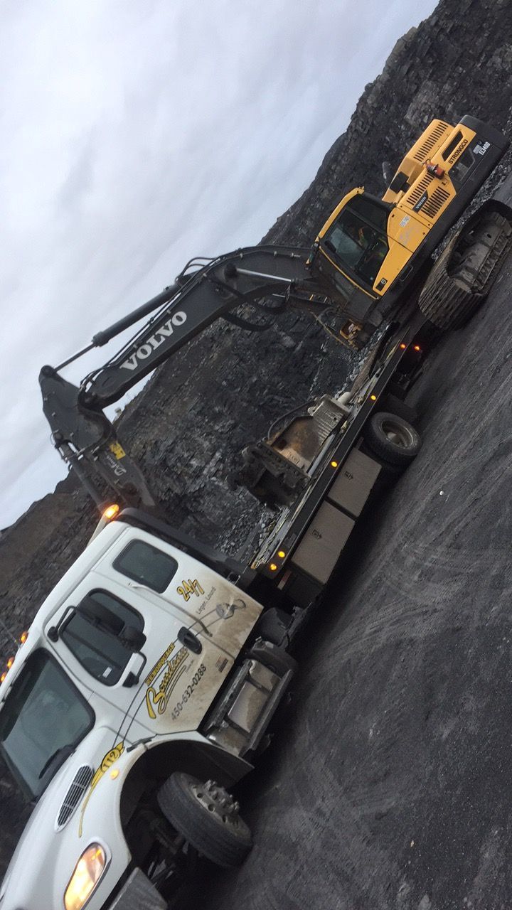 Une dépanneuse blanche transporte une excavatrice Volvo jaune sur une surface graveleuse sous un ciel nuageux.