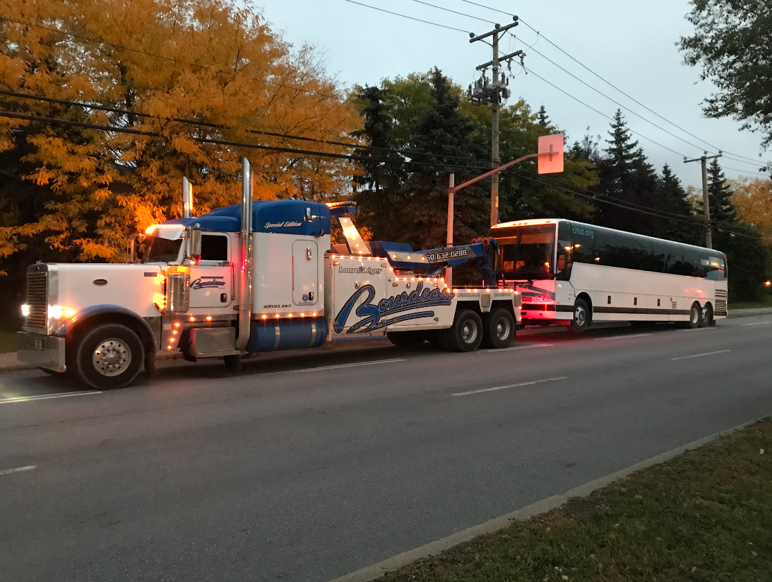Une grosse dépanneuse tracte un bus blanc sur une route au crépuscule. Le camion est bleu et blanc, avec le nom de l'entreprise inscrit sur le côté.