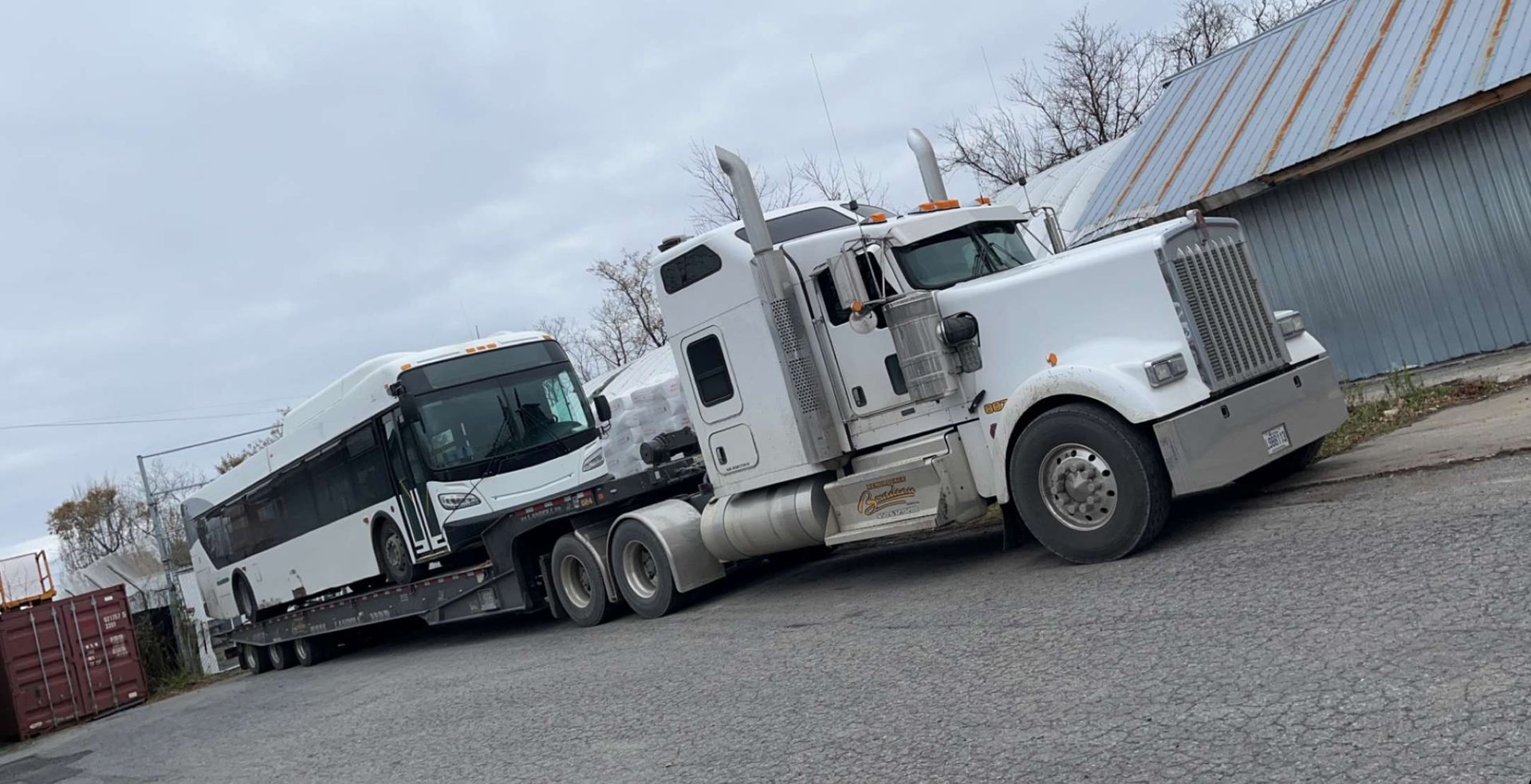 Un semi-camion blanc tirant un bus blanc sur une remorque, garé à l'extérieur près d'un bâtiment sous un ciel nuageux.