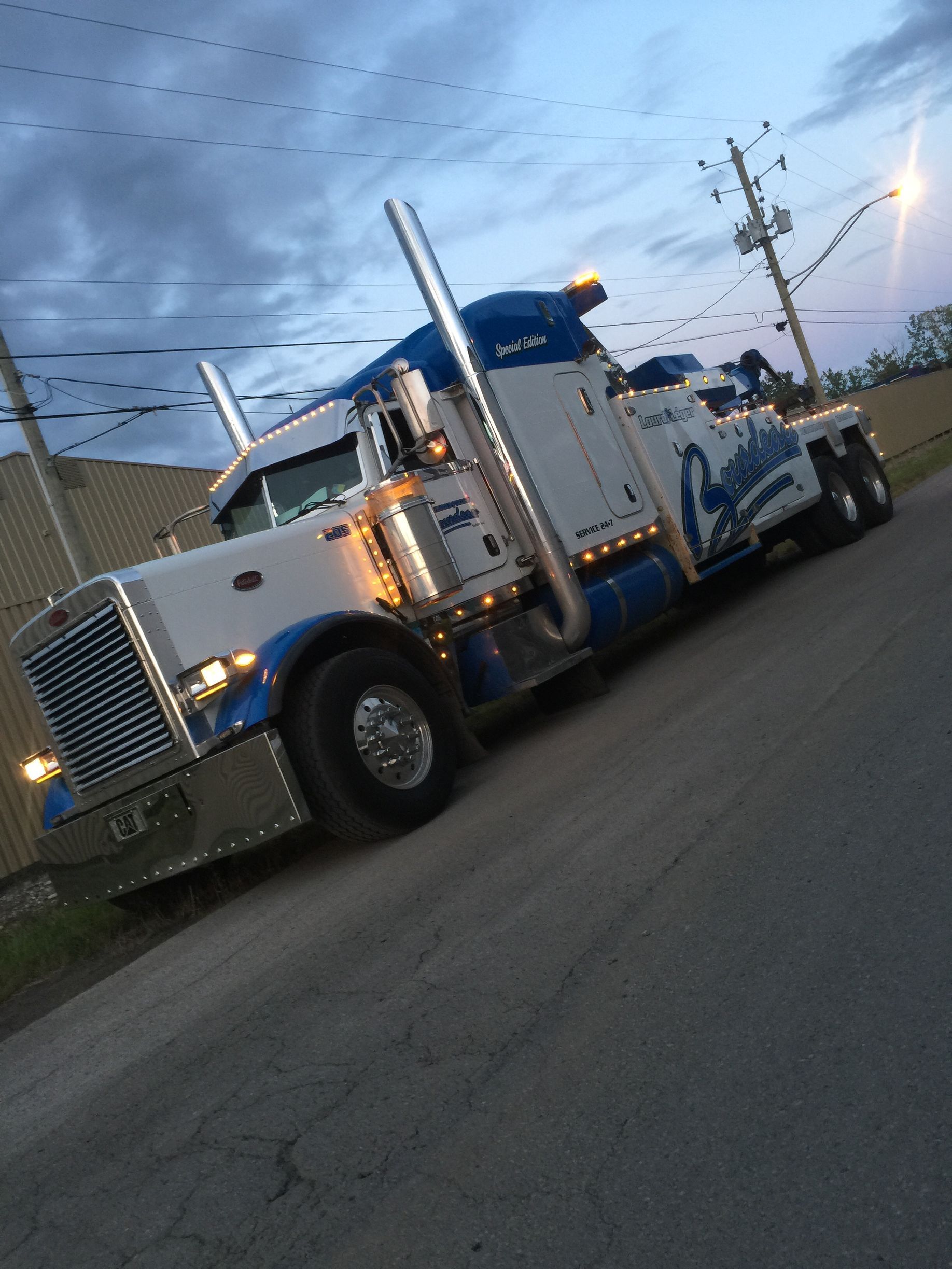 Dépanneuse blanche et bleue garée au bord d'une route au crépuscule. Le véhicule est incliné et arbore une peinture personnalisée.