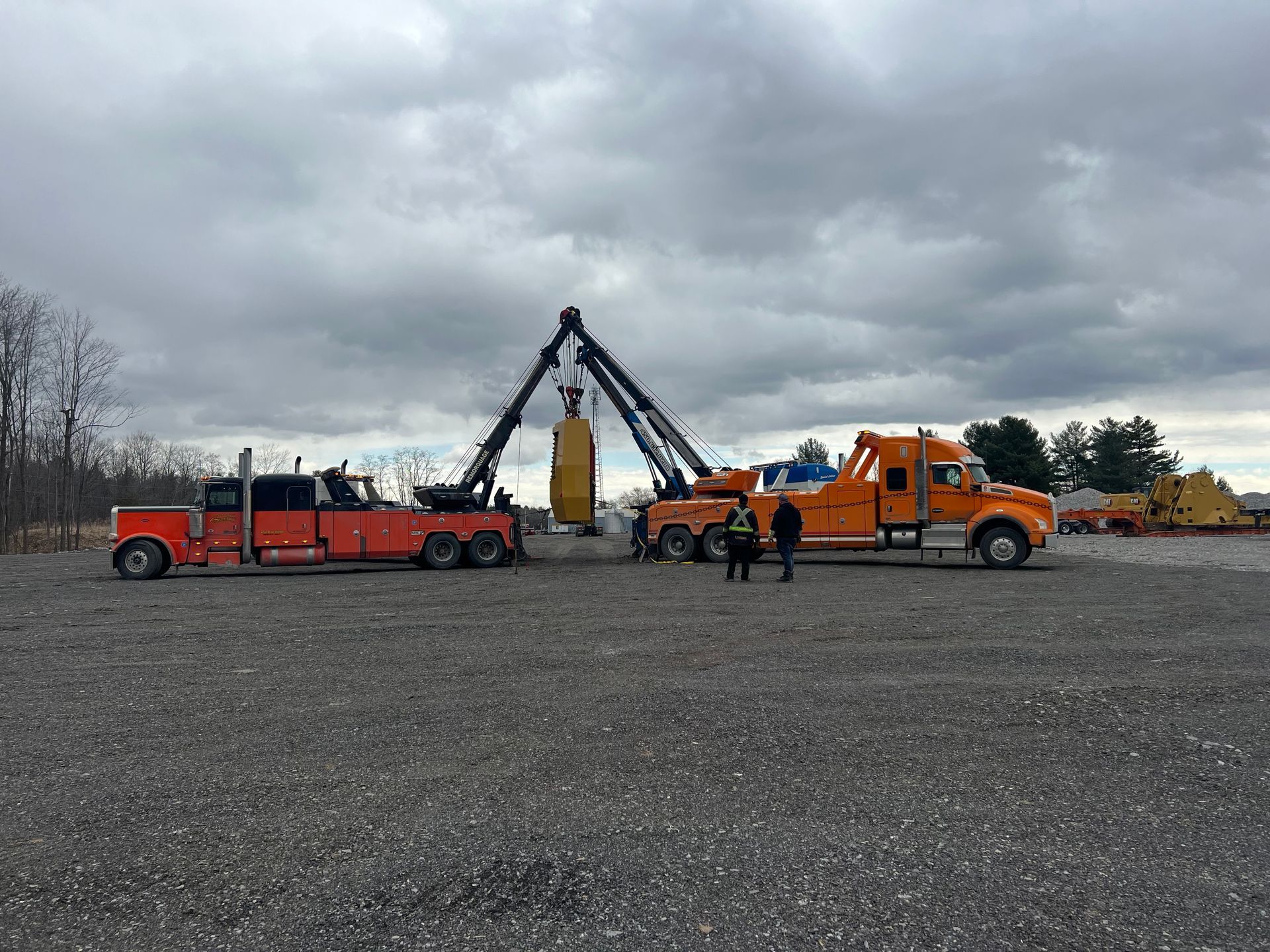 Deux dépanneuses orange soulèvent un grand objet jaune à l'extérieur, par temps nuageux. Deux personnes se tiennent près de l'objet.