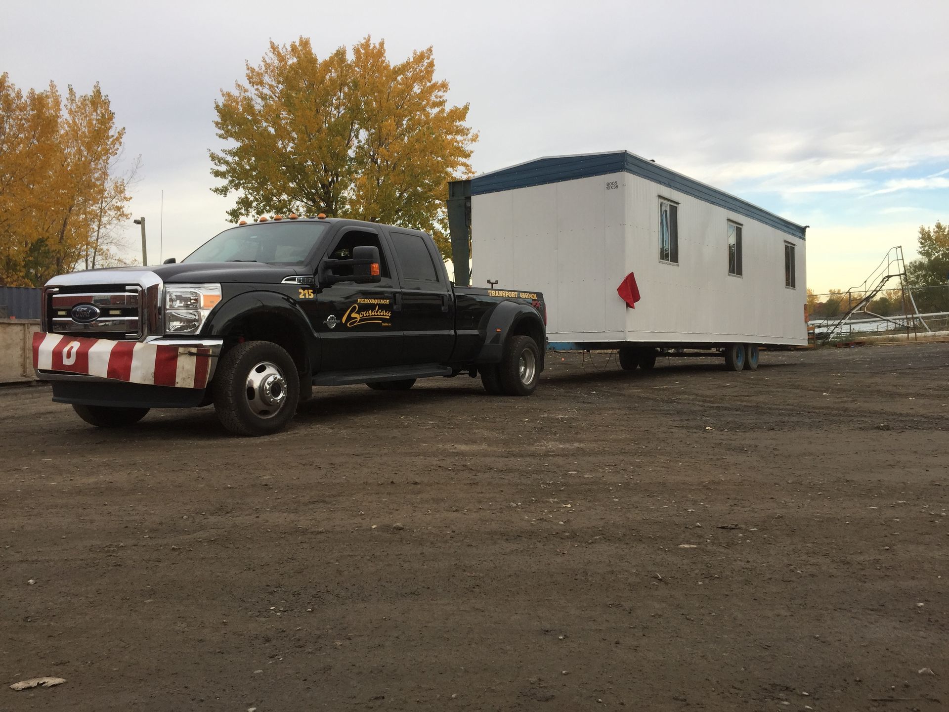 Camionnette noire remorquant un bâtiment portable blanc et bleu sur un terrain en terre, sous un ciel nuageux.
