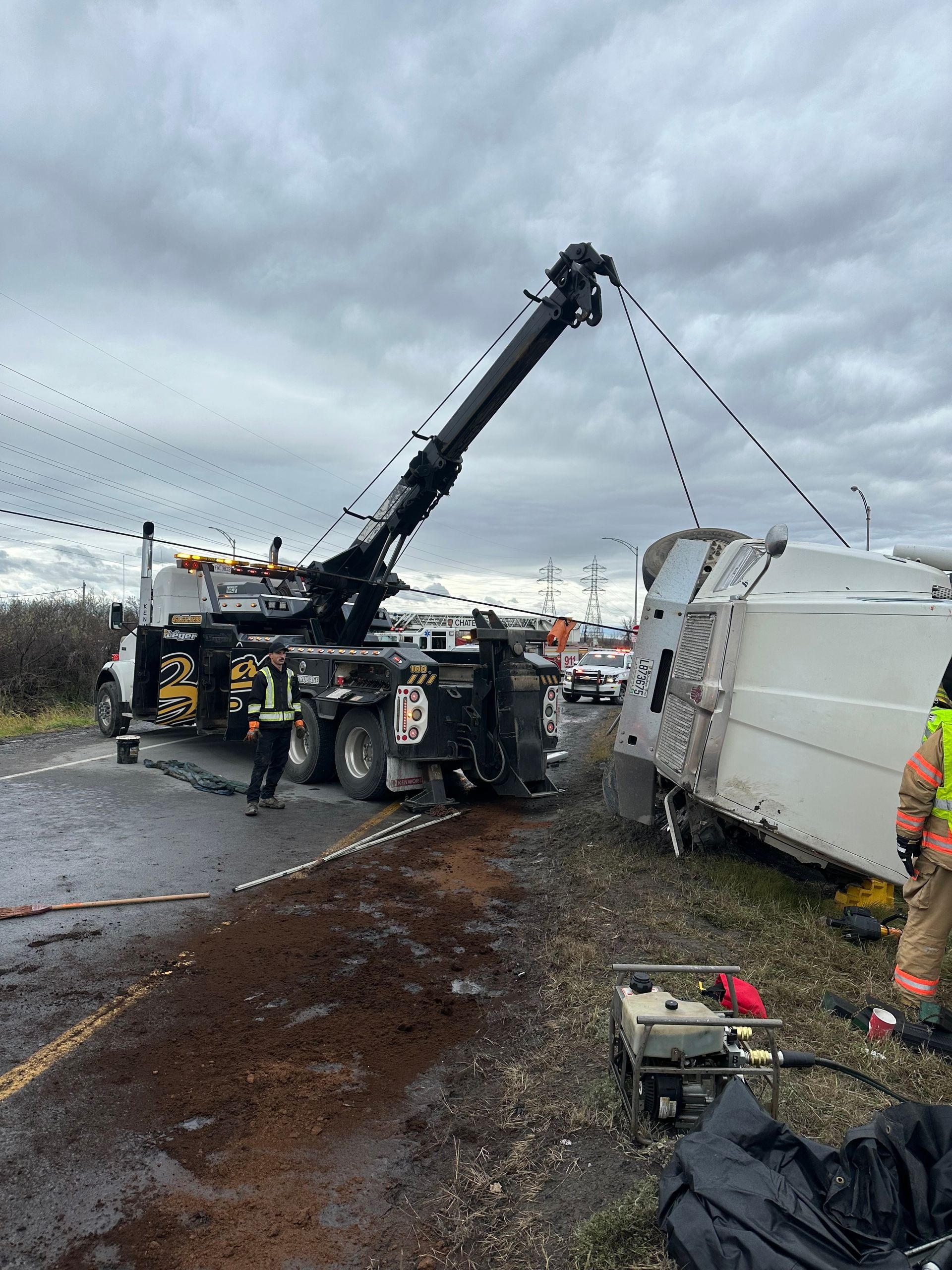 Une dépanneuse redresse un véhicule blanc renversé sur le bord de la route, sous un ciel nuageux. Les secours sont présents.