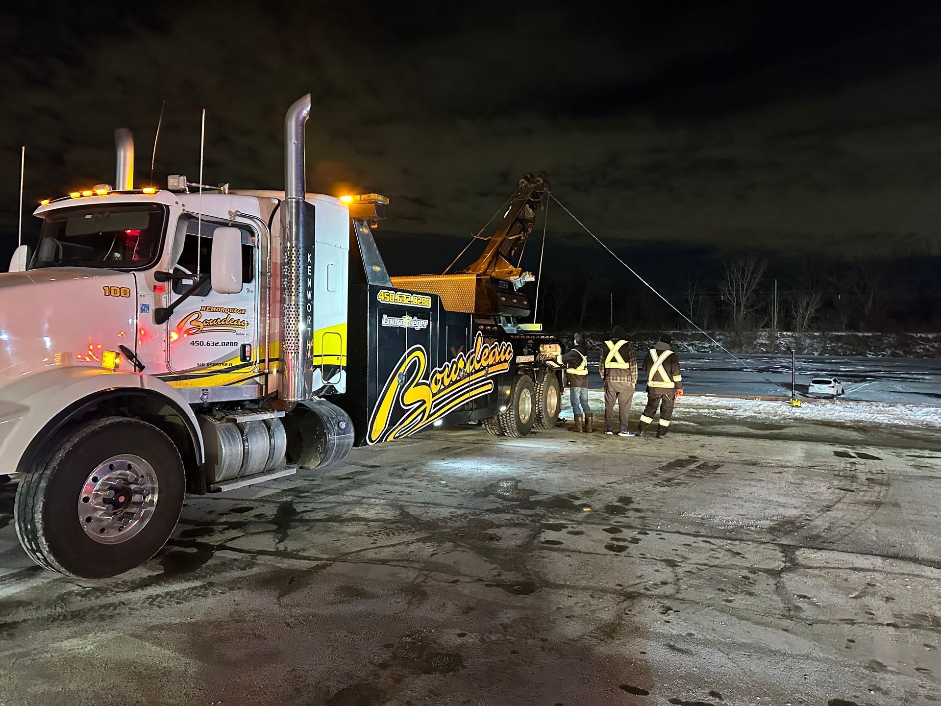 Une dépanneuse la nuit avec plusieurs personnes portant des gilets de sécurité à proximité d'un véhicule endommagé.