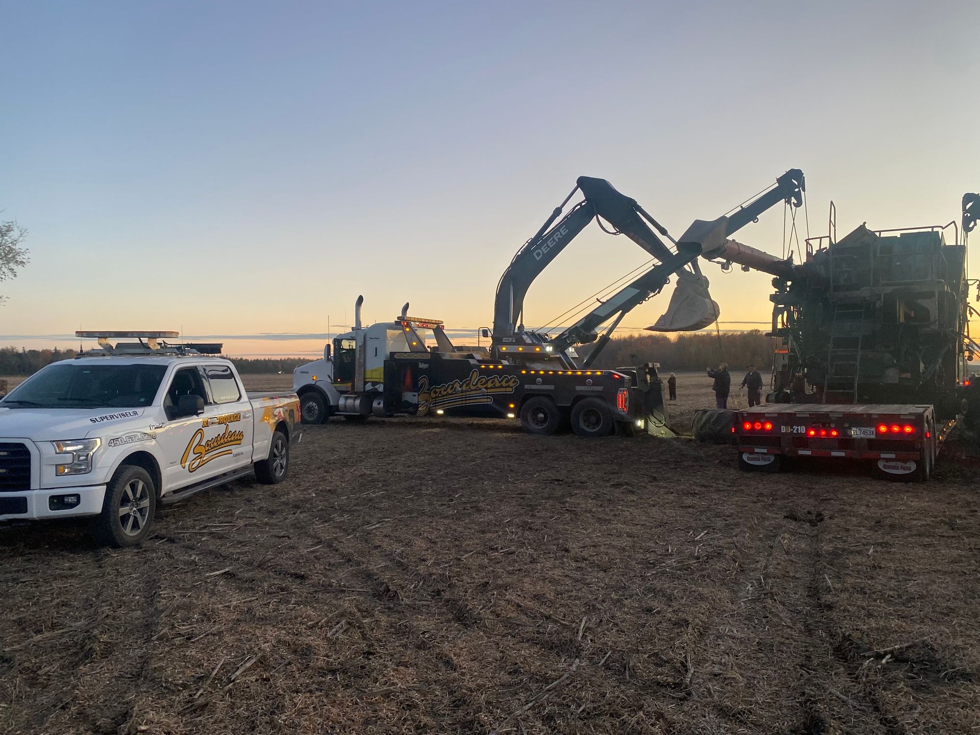 Une dépanneuse et une excavatrice s'efforcent de soulever une grosse machine agricole dans un champ au crépuscule. Un camion de service est garé à proximité.