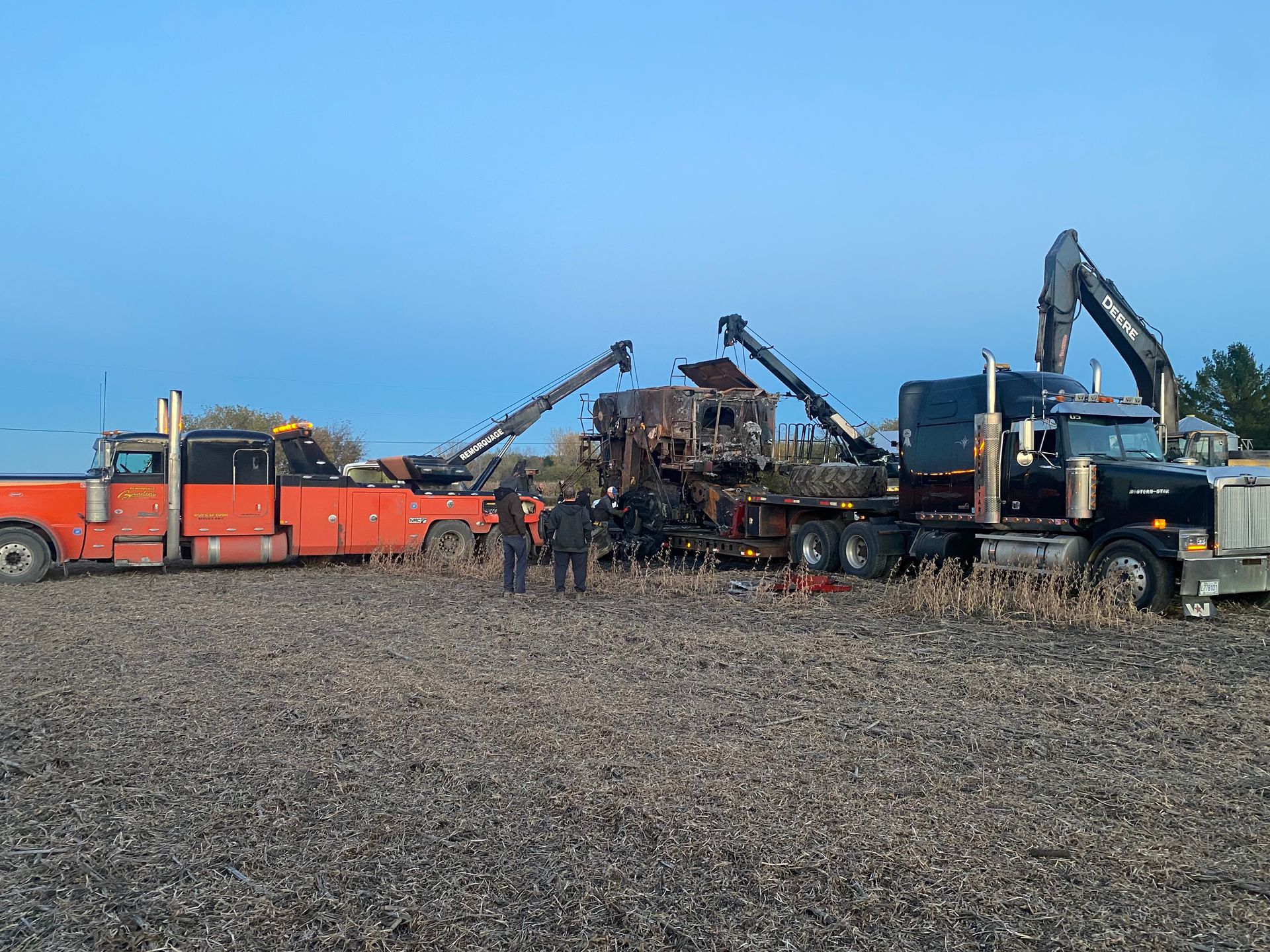 Un semi-remorque noir et une dépanneuse rouge équipée de grues chargent une machine calcinée dans un champ. Plusieurs personnes se tiennent à proximité.