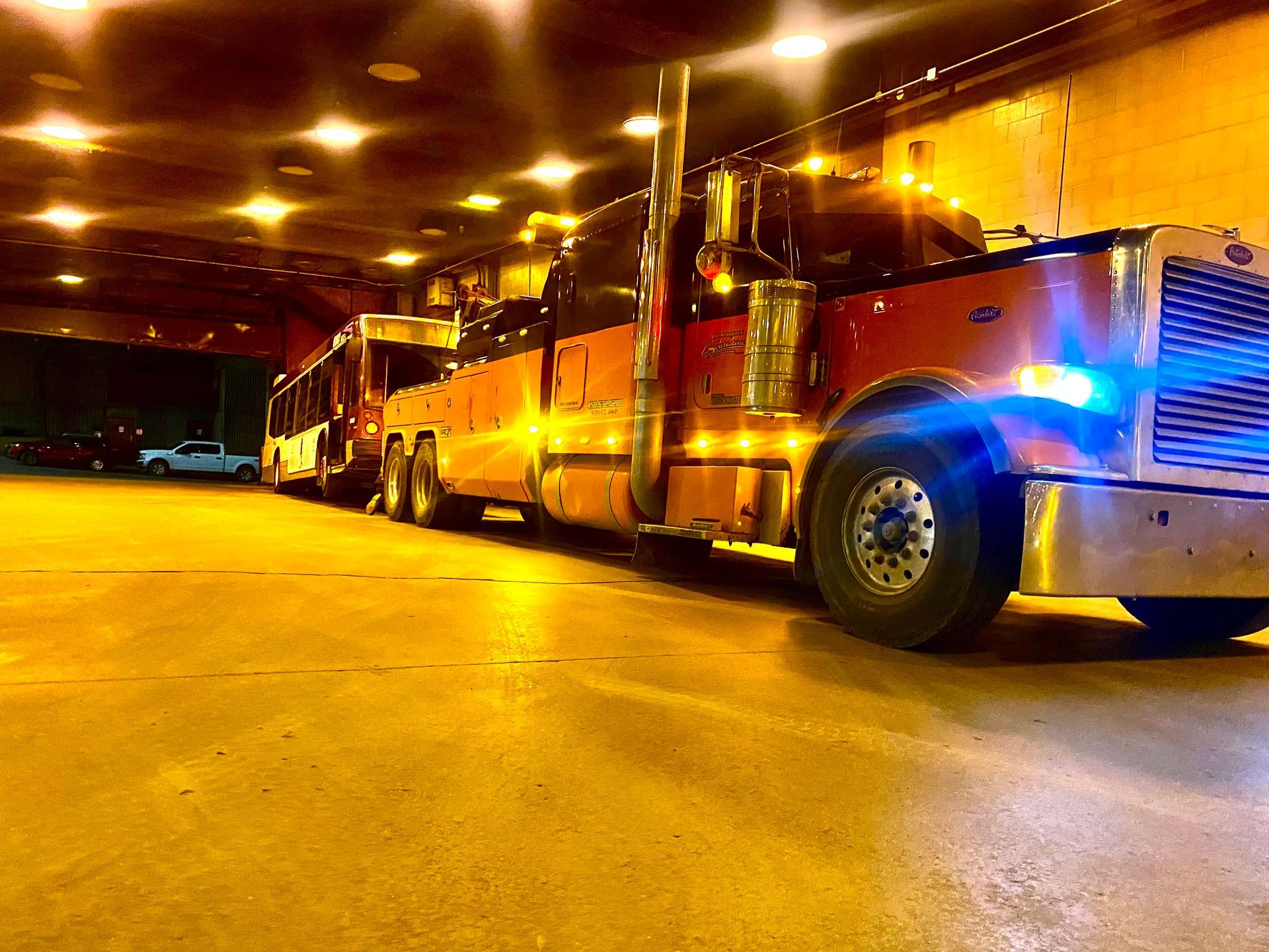 Une grosse dépanneuse illuminée par des lumières vives est garée dans un garage ou un tunnel. Sa peinture rouge et orange scintille, avec des lumières bleues allumées.