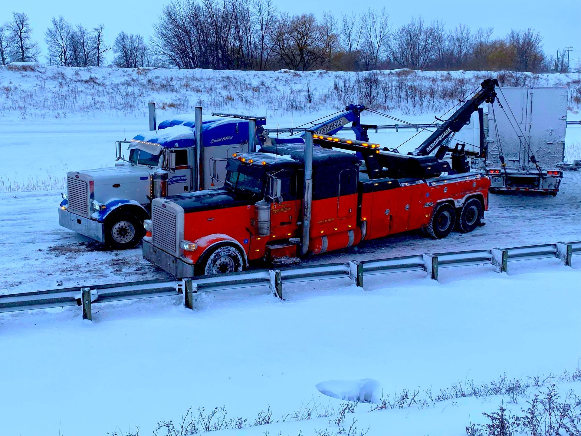Deux dépanneuses, une rouge et une bleue, stationnées sur le bord d'une route enneigée, probablement pour aider une semi-remorque en panne.