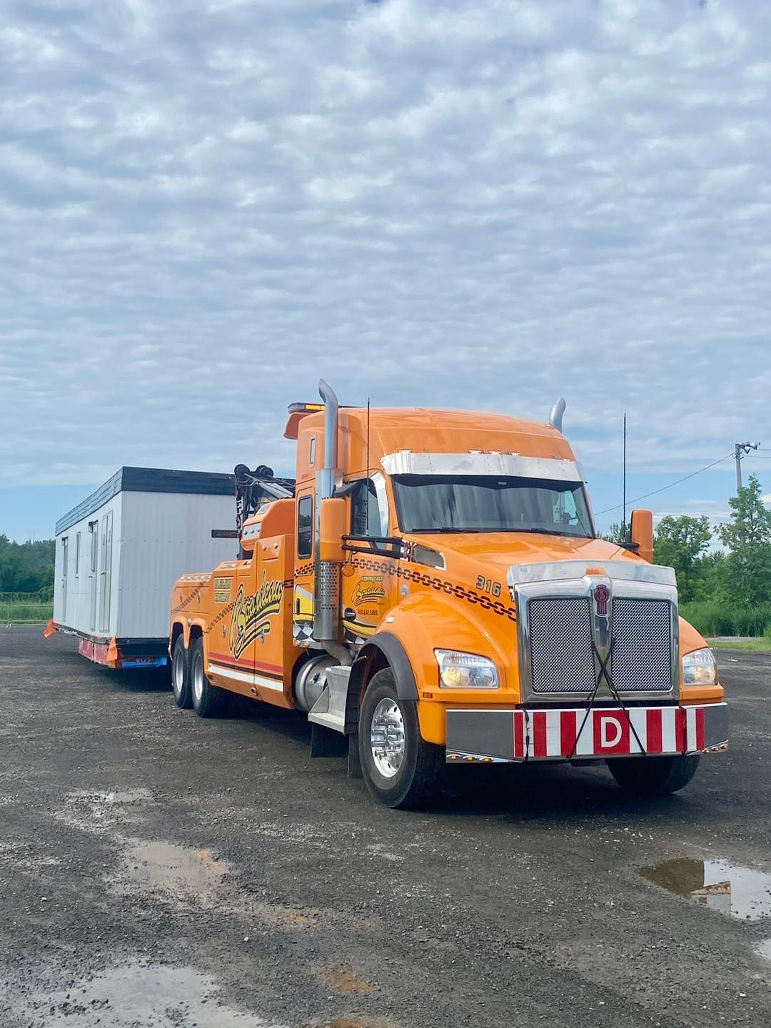 Dépanneuse orange transportant un conteneur blanc sur un terrain gris sous un ciel nuageux.