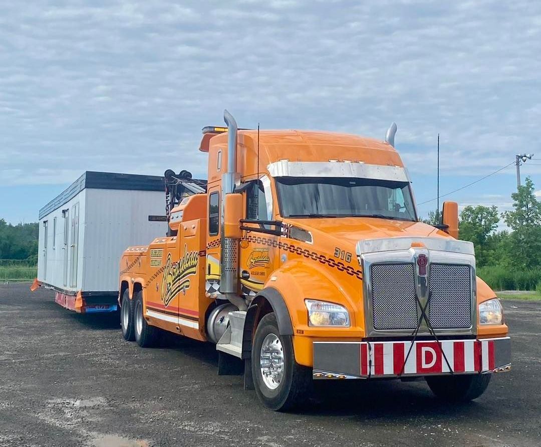 Dépanneuse orange tirant un bâtiment blanc et gris par temps nuageux. Le camion est doté d'une calandre chromée brillante et d'un pare-chocs en forme de D.