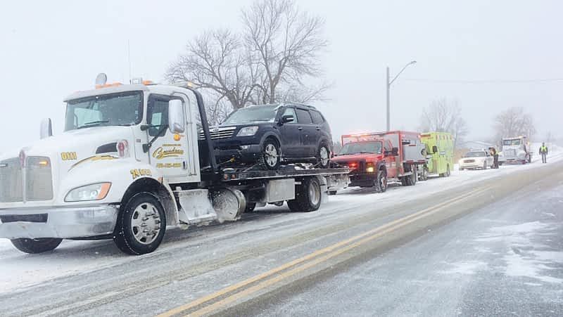 Une dépanneuse avec une voiture sur son plateau, suivie de véhicules d'urgence, alignés sur une route enneigée. Scène hivernale.