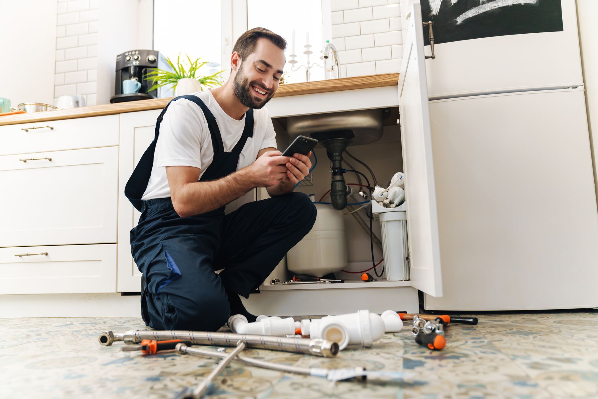 Repair worker kneels under open sink, checking phone, plumbing tools on floor.