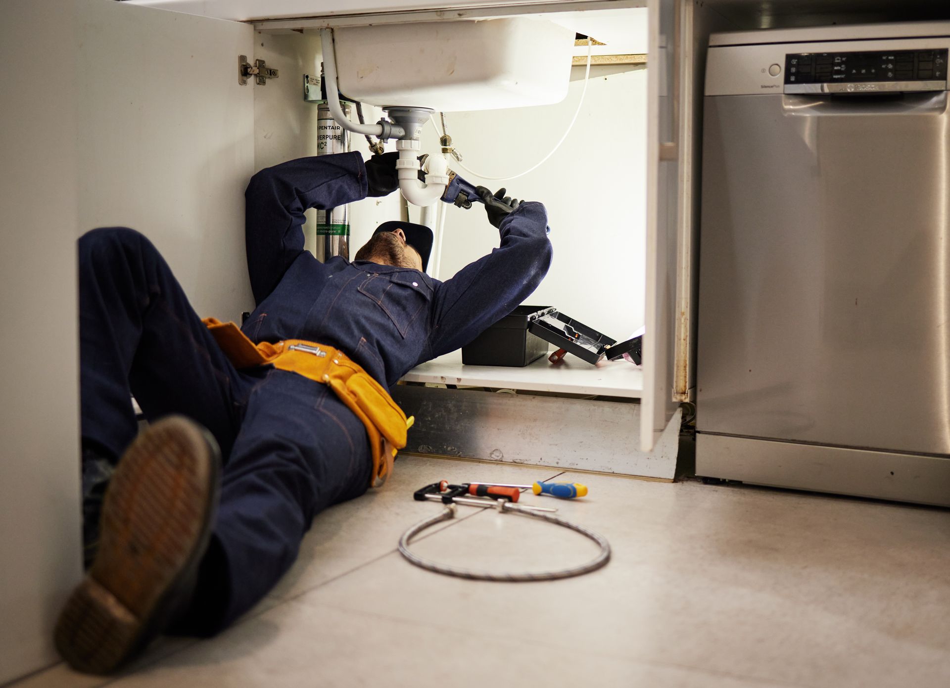 Plumber lying on the floor repairing pipes beneath a kitchen sink, with tools scattered nearby.