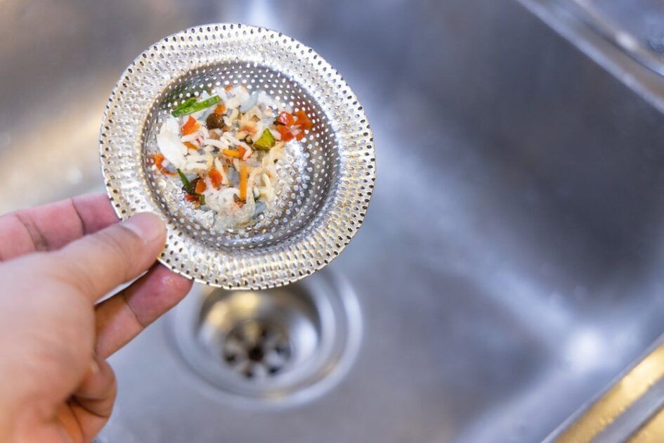 A person is holding a strainer with food in it over a sink drain.