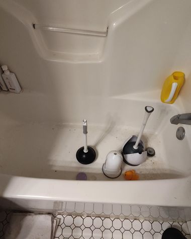 Bathtub with a toilet plunger, toilet brush, and cleaning supplies scattered inside.