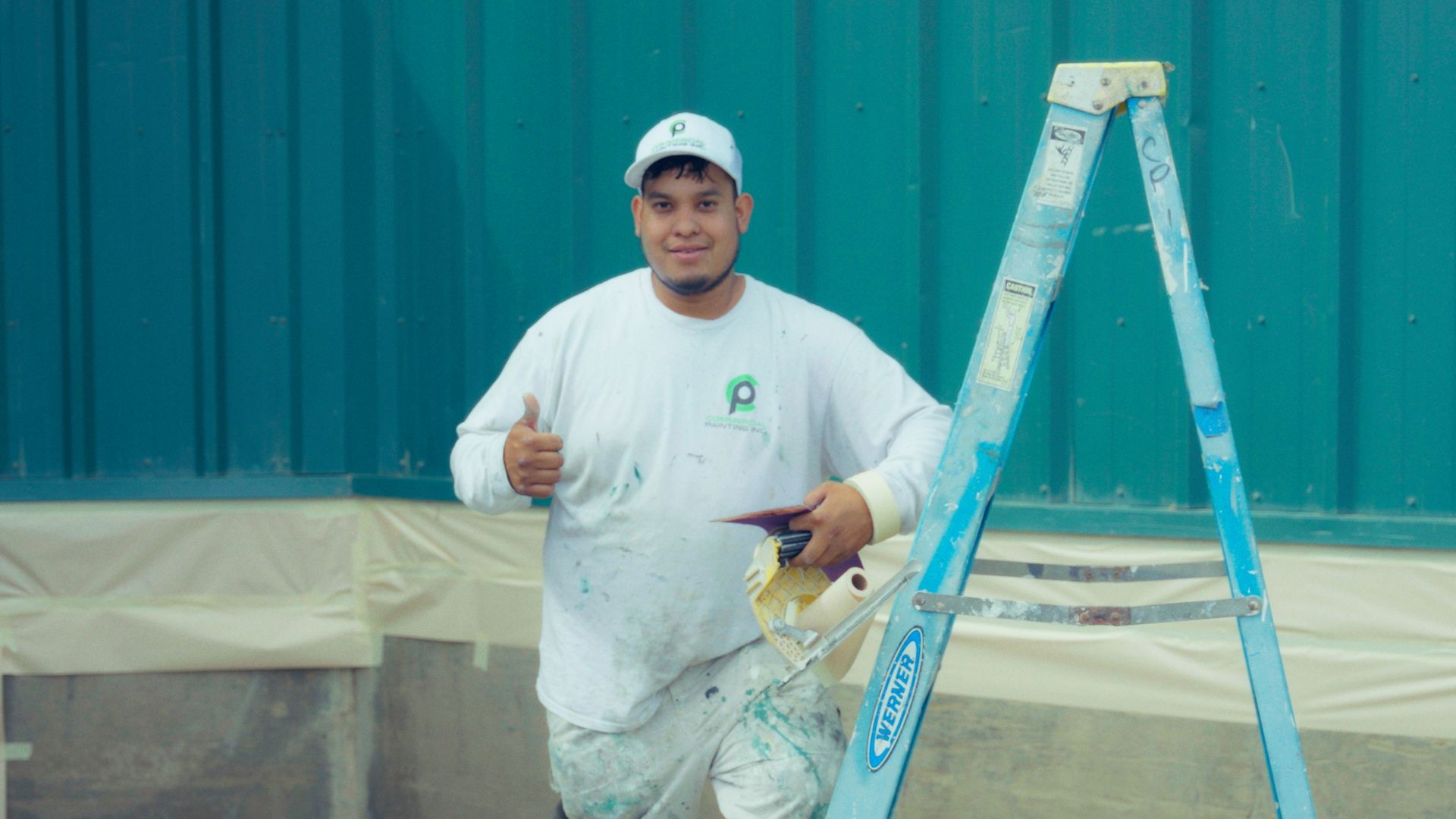 Painter in white clothing and cap gives a thumbs-up next to a ladder in front of a teal wall.