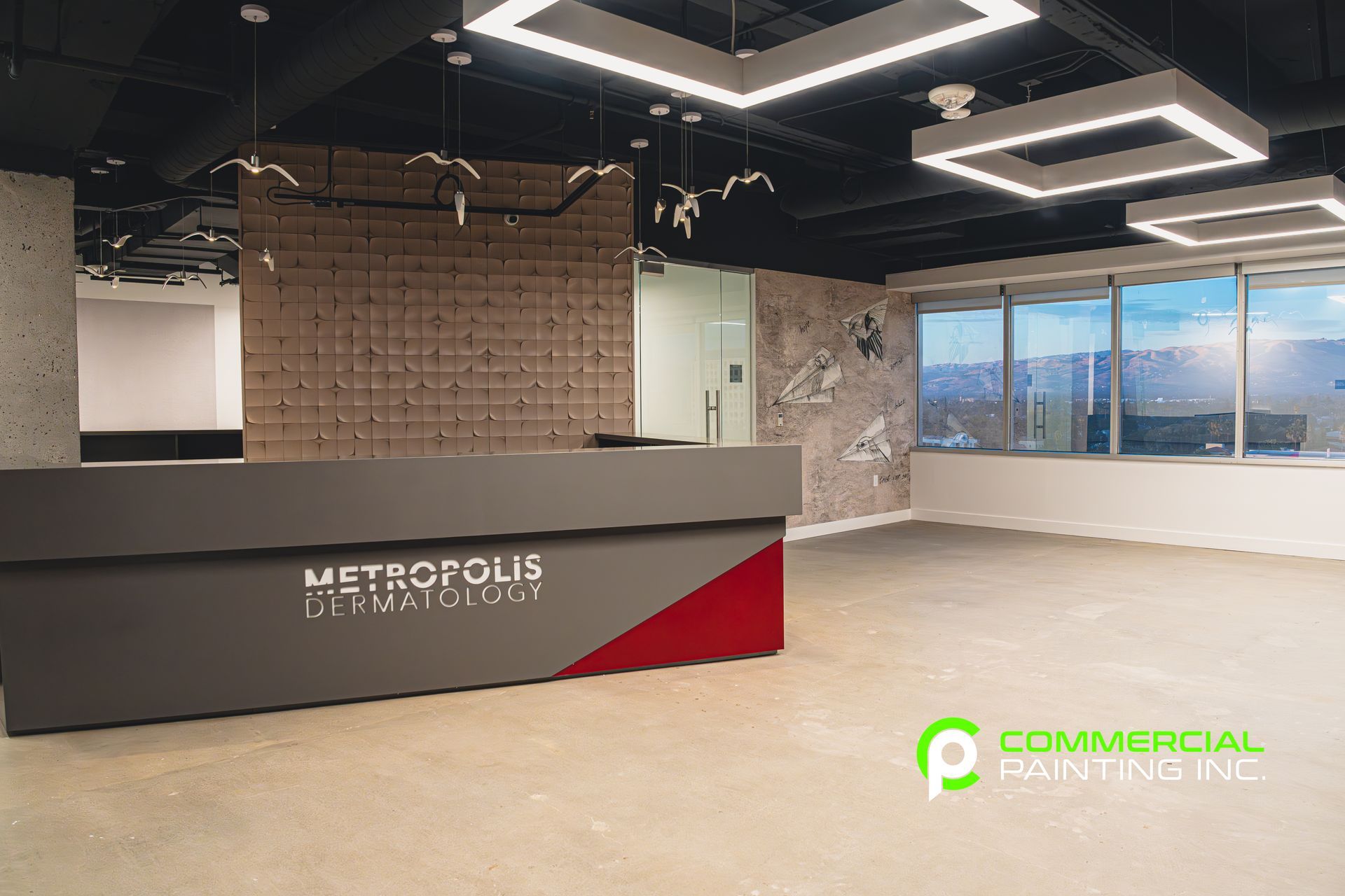 Modern office reception area with a gray and red desk, wood-paneled wall, and square ceiling lights. The windows offer a cityscape view.