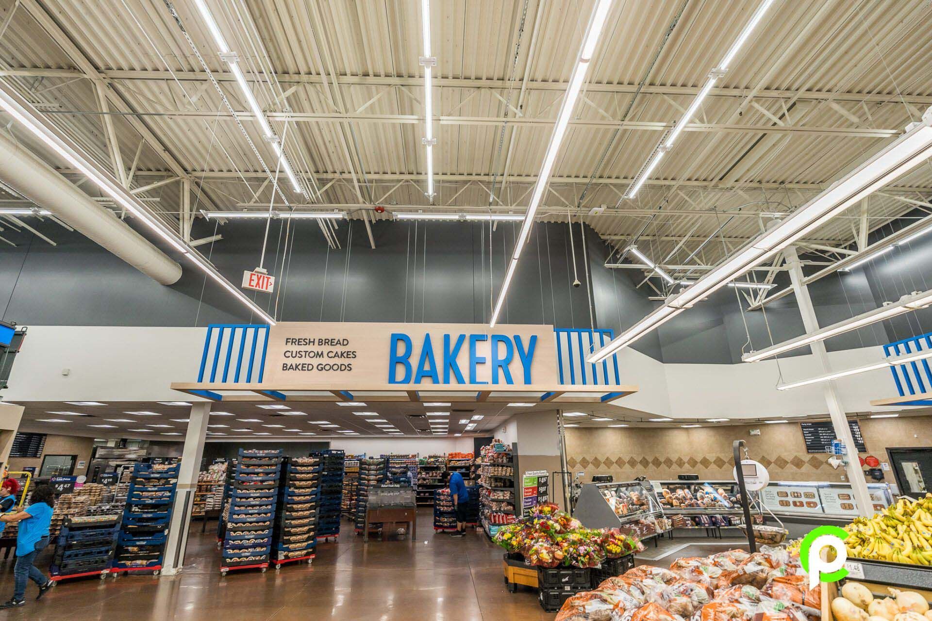 Inside a supermarket, a sign for the bakery is visible. Blue and white bakery sign. People are shopping, and shelves are stocked.