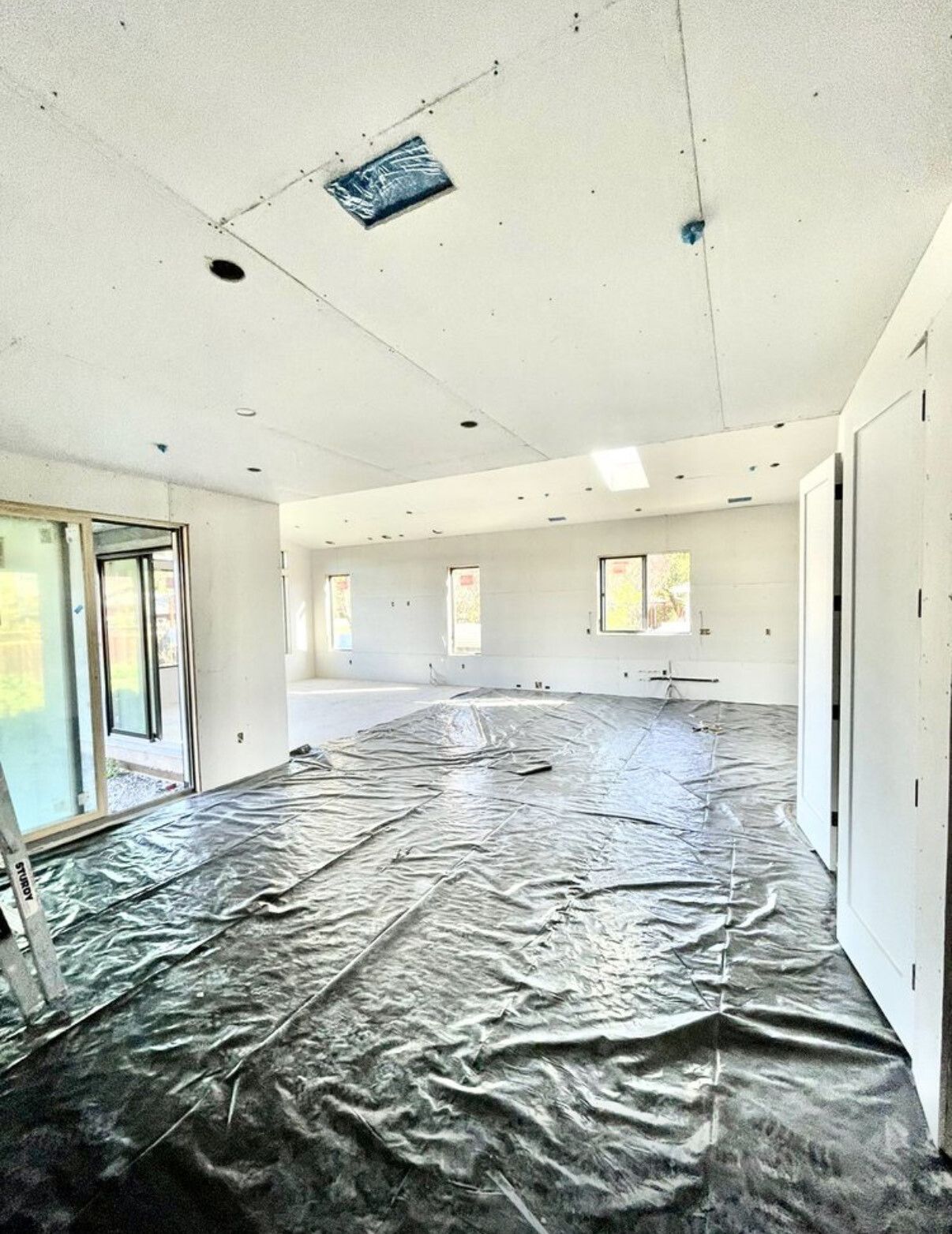 Interior of a room under construction with protective sheeting on the floor. Drywall, windows, and doors are visible.