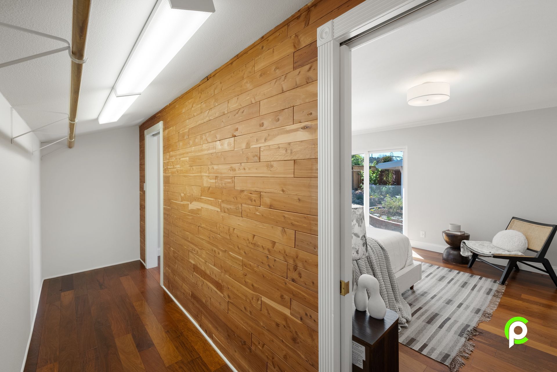 Hallway with wood accent wall, leading to bedroom with bed, chair, and outdoor view.