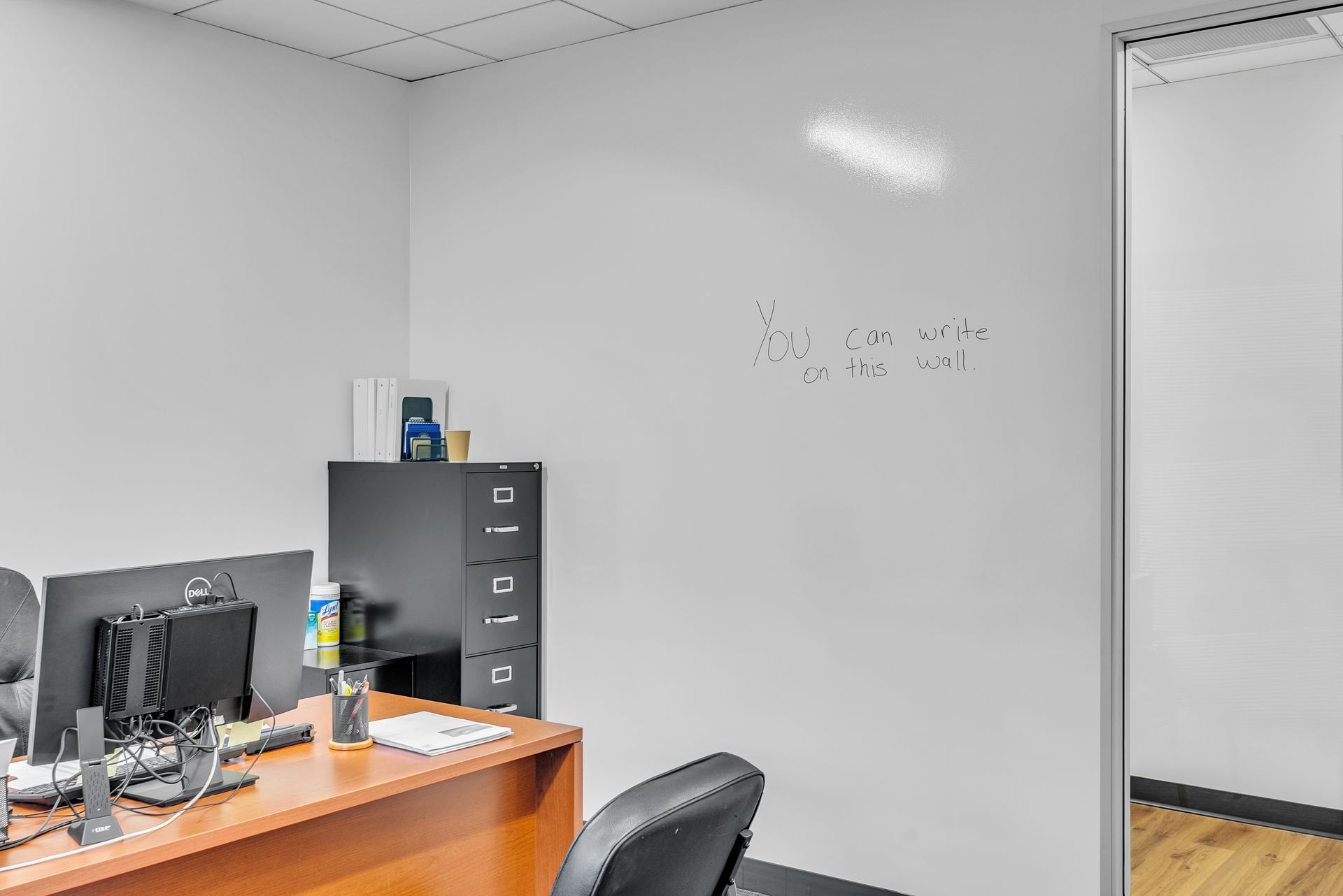 Office corner: desk with computer monitors, black filing cabinet, whiteboard with writing, and an open doorway.