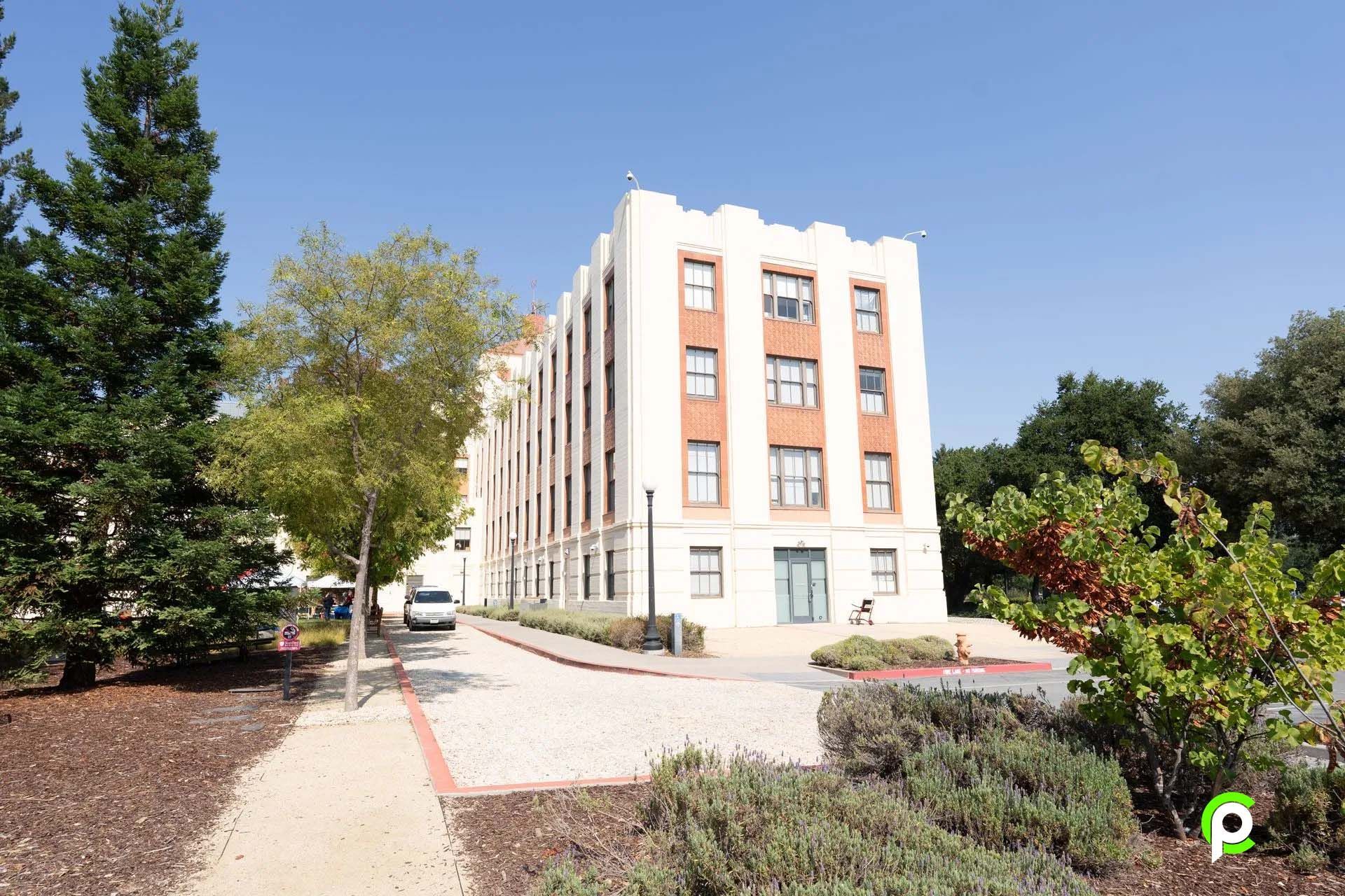 A large white building with a lot of windows is surrounded by trees and bushes.