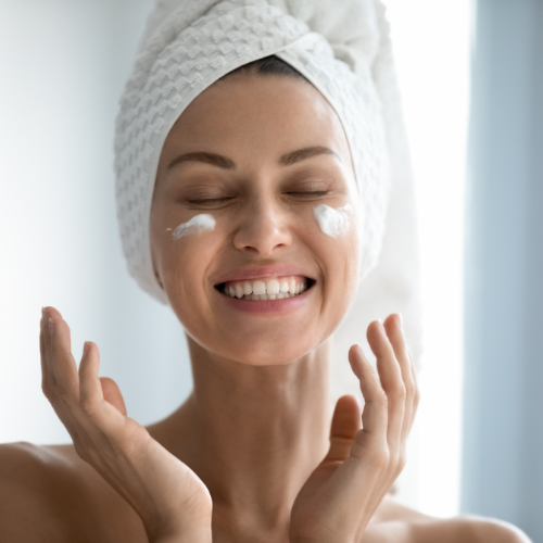 Eine Frau mit einem Handtuch um den Kopf lächelt und cremt ihr Gesicht ein.