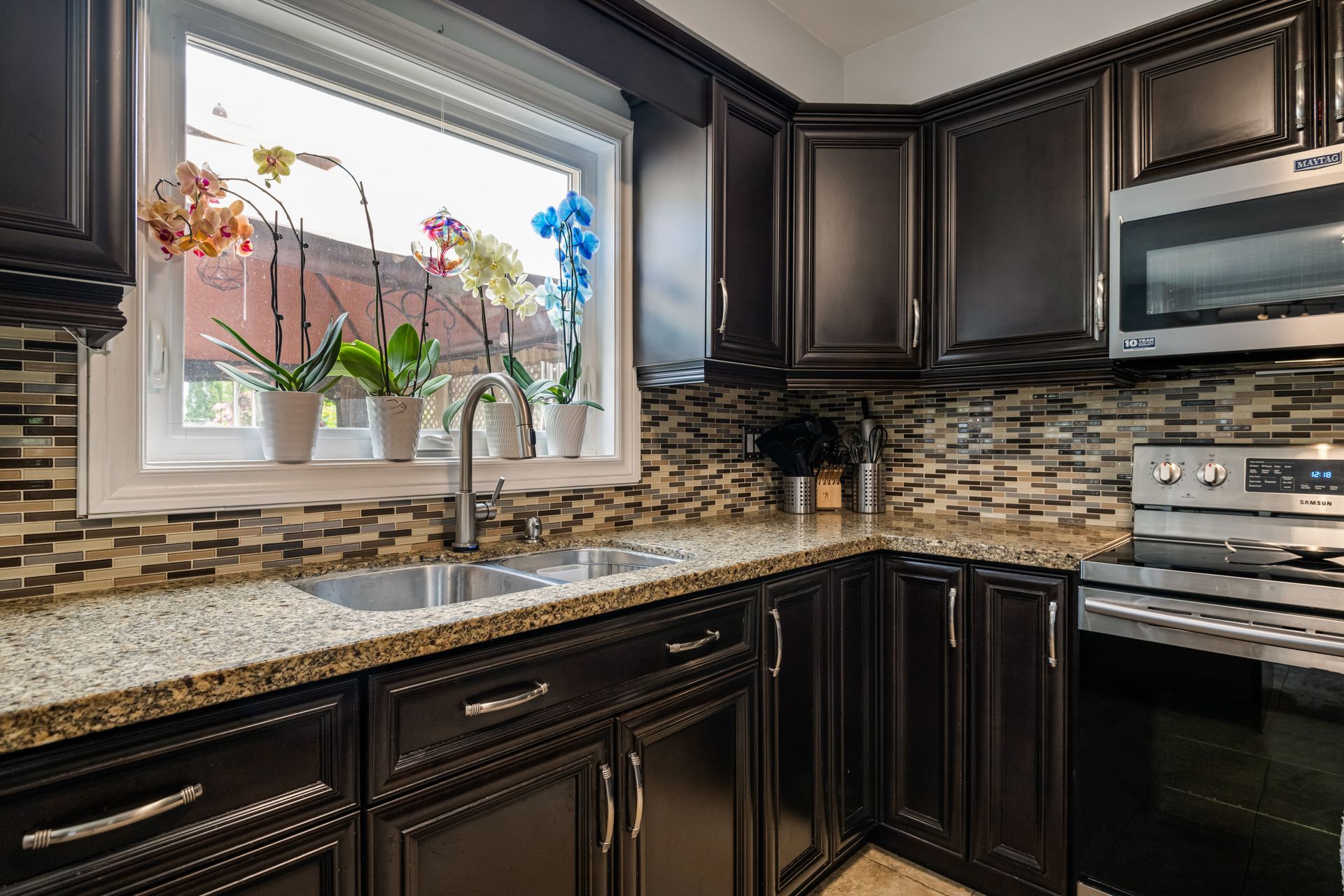 A kitchen with black cabinets , granite counter tops , stainless steel appliances and a large window.