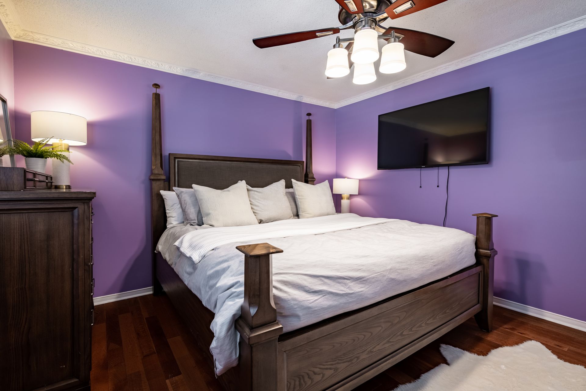 A bedroom with purple walls , a bed , a television and a ceiling fan.
