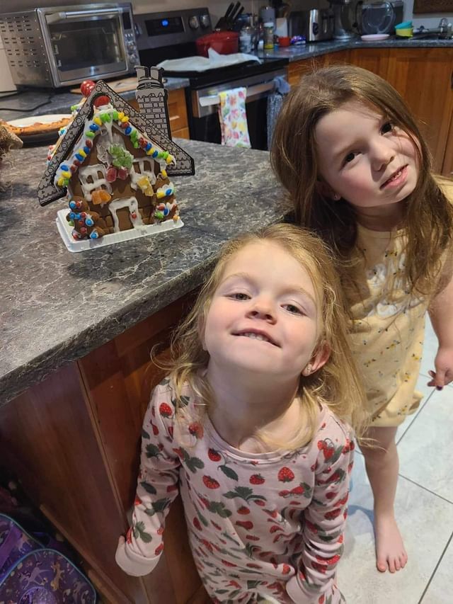 Two little girls are standing next to a gingerbread house in a kitchen.