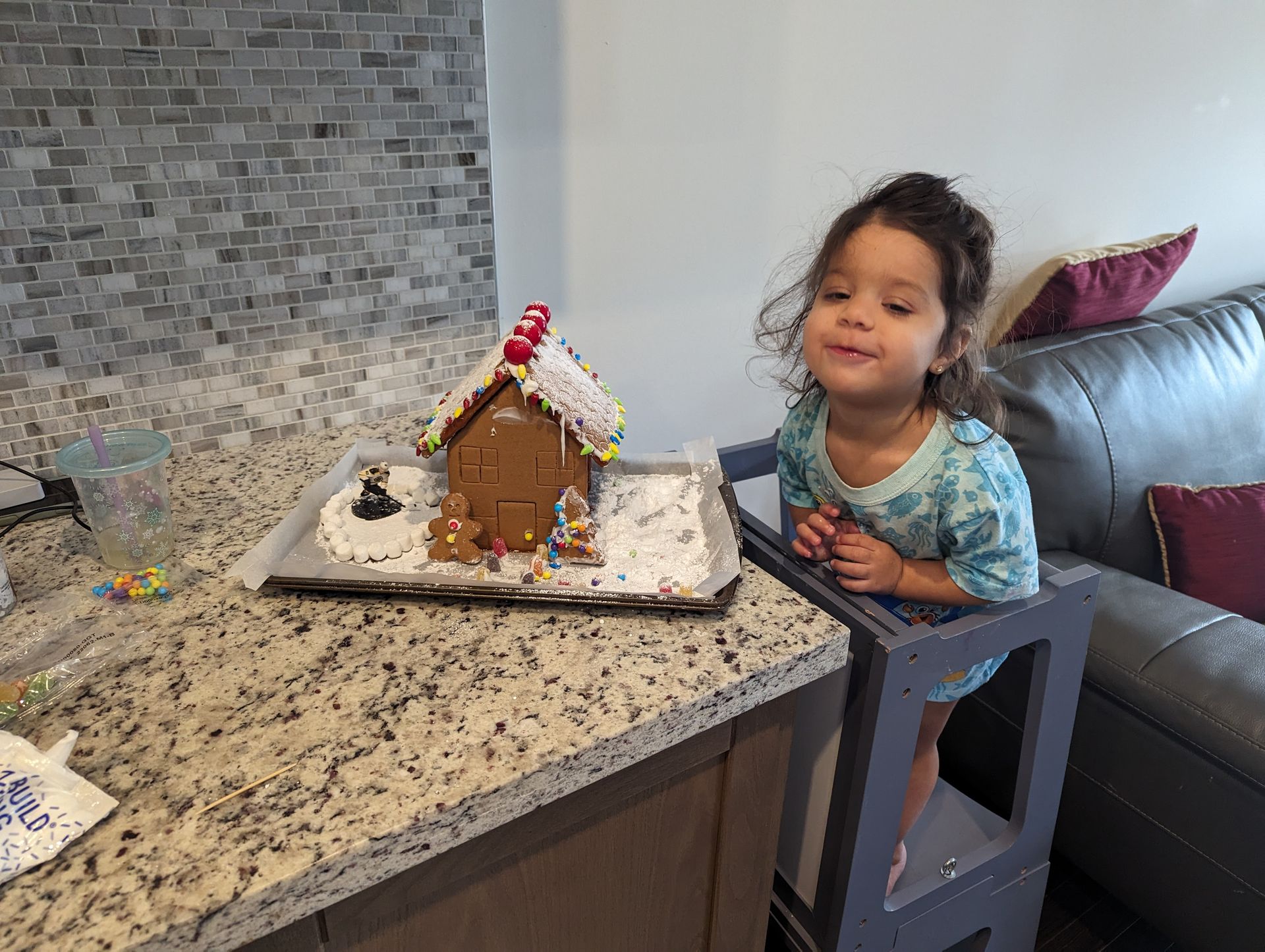 A little girl is sitting on a stool in front of a gingerbread house.